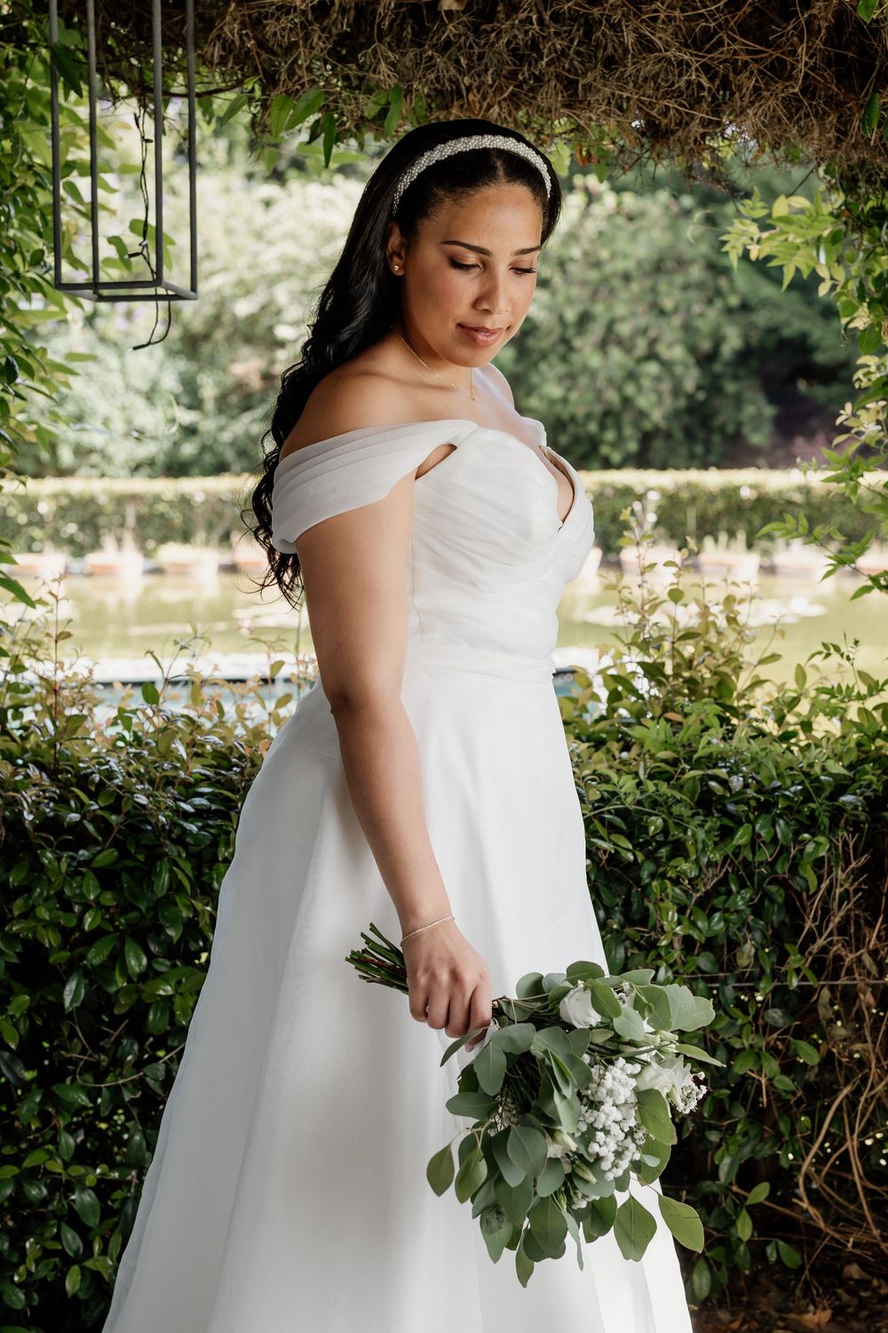 A person in a white off-shoulder wedding dress holds a green eucalyptus bouquet in an outdoor garden setting.