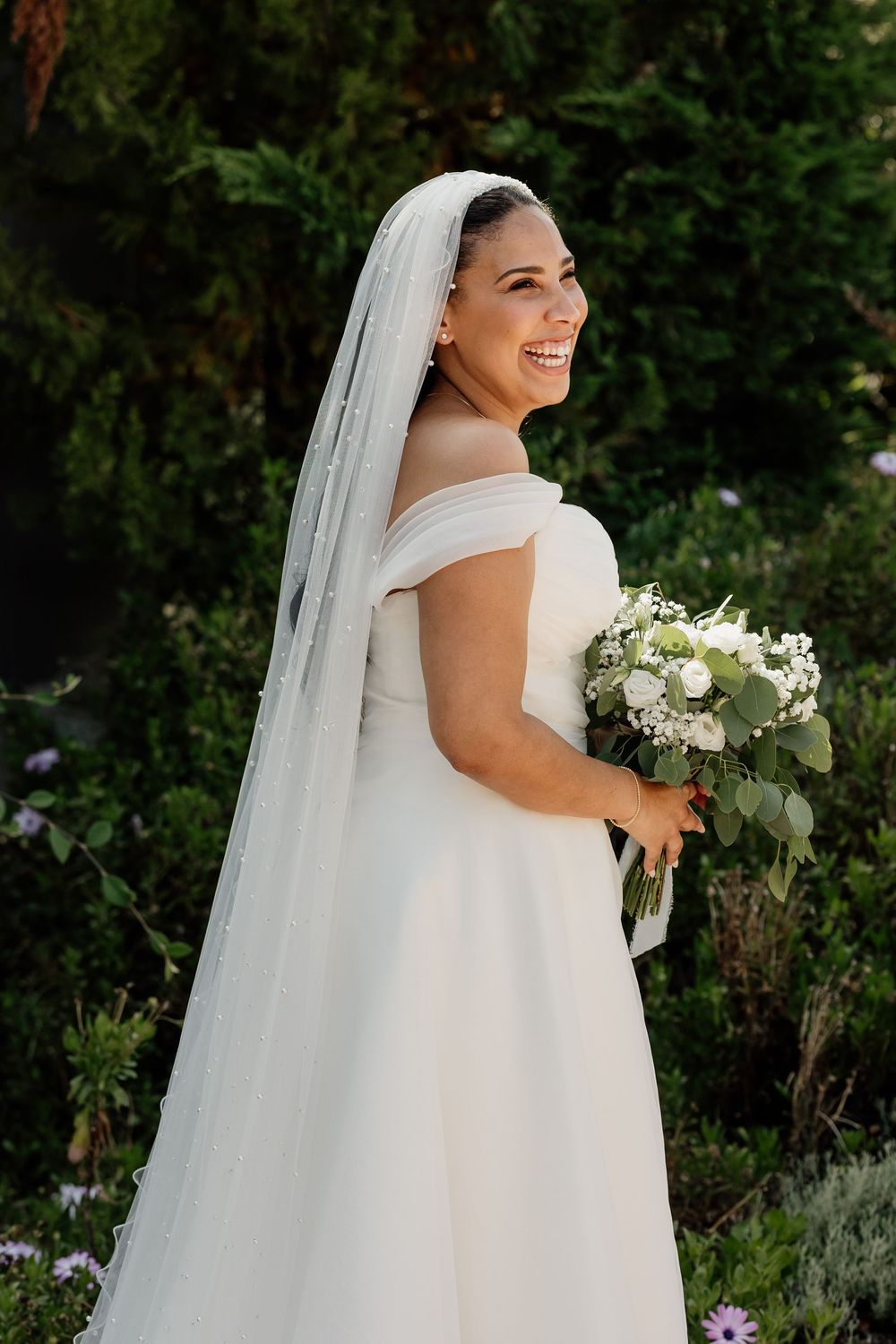 A bride in an elegant off-shoulder white wedding gown and veil poses outdoors against a lush garden backdrop.