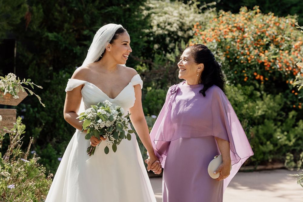 A wedding party in lavender and white dresses poses together in a garden setting with vibrant orange flowers.