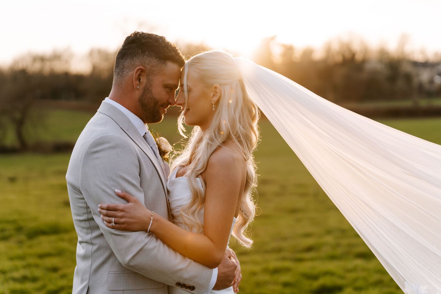 A romantic wedding couple embraces in a sunlit field while a long white veil flows in the breeze.