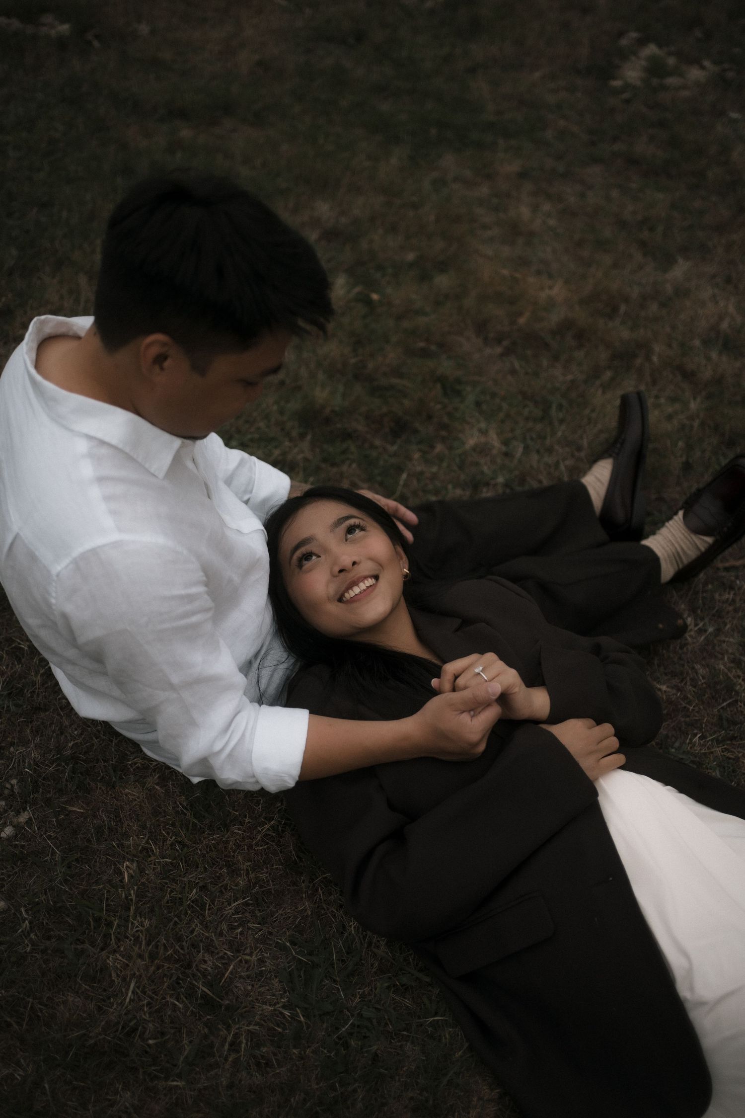 A couple in formal attire lying on grass and sharing playful moments during a photoshoot after their proposal in Yarra Glen