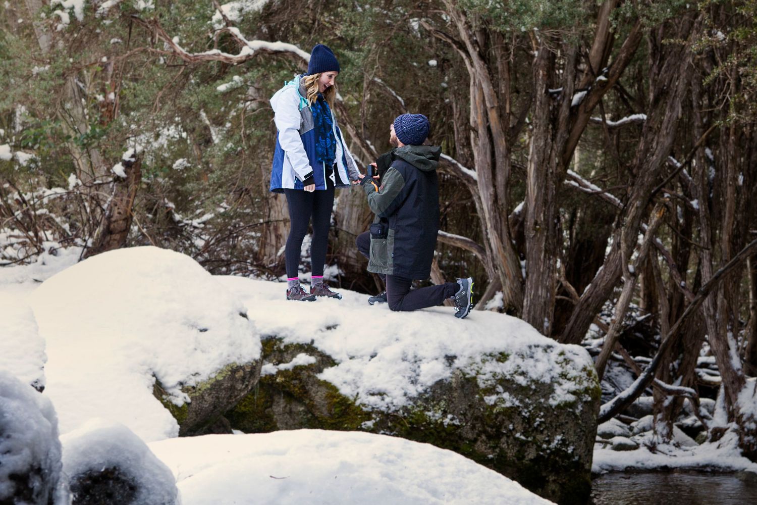 A snowy winter hiking trail winds through a forest. Hiker stops and proposes to his girlfriend, her face excited
