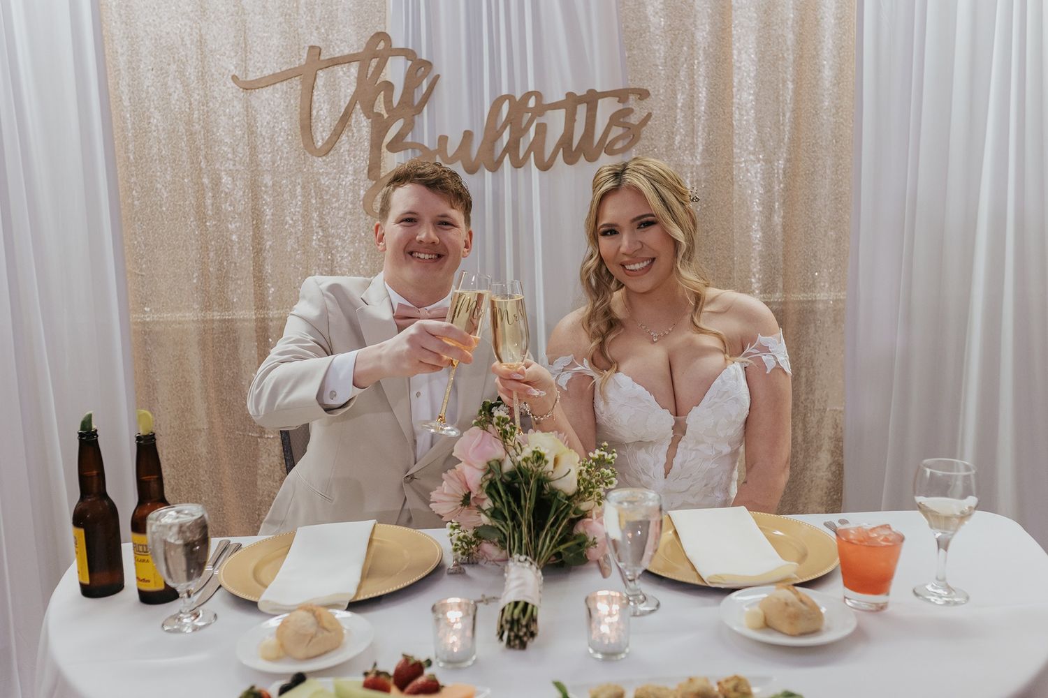 Wedding guests raise champagne glasses in a toast at an elegant reception dinner with white tablecloths and floral decor.