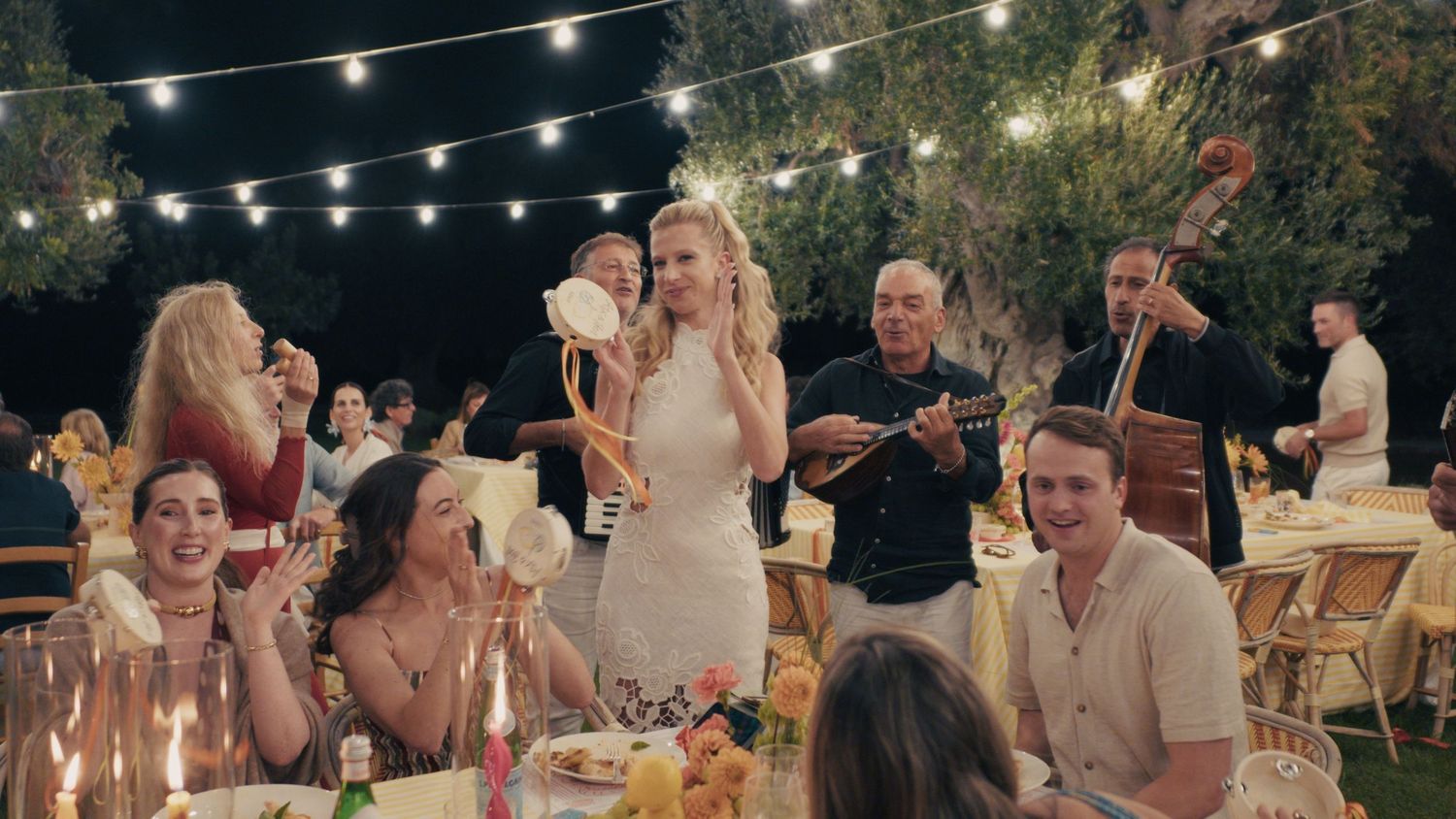 Outdoor wedding reception at night with string lights, guests dining, bride in white dress, and live band performing.