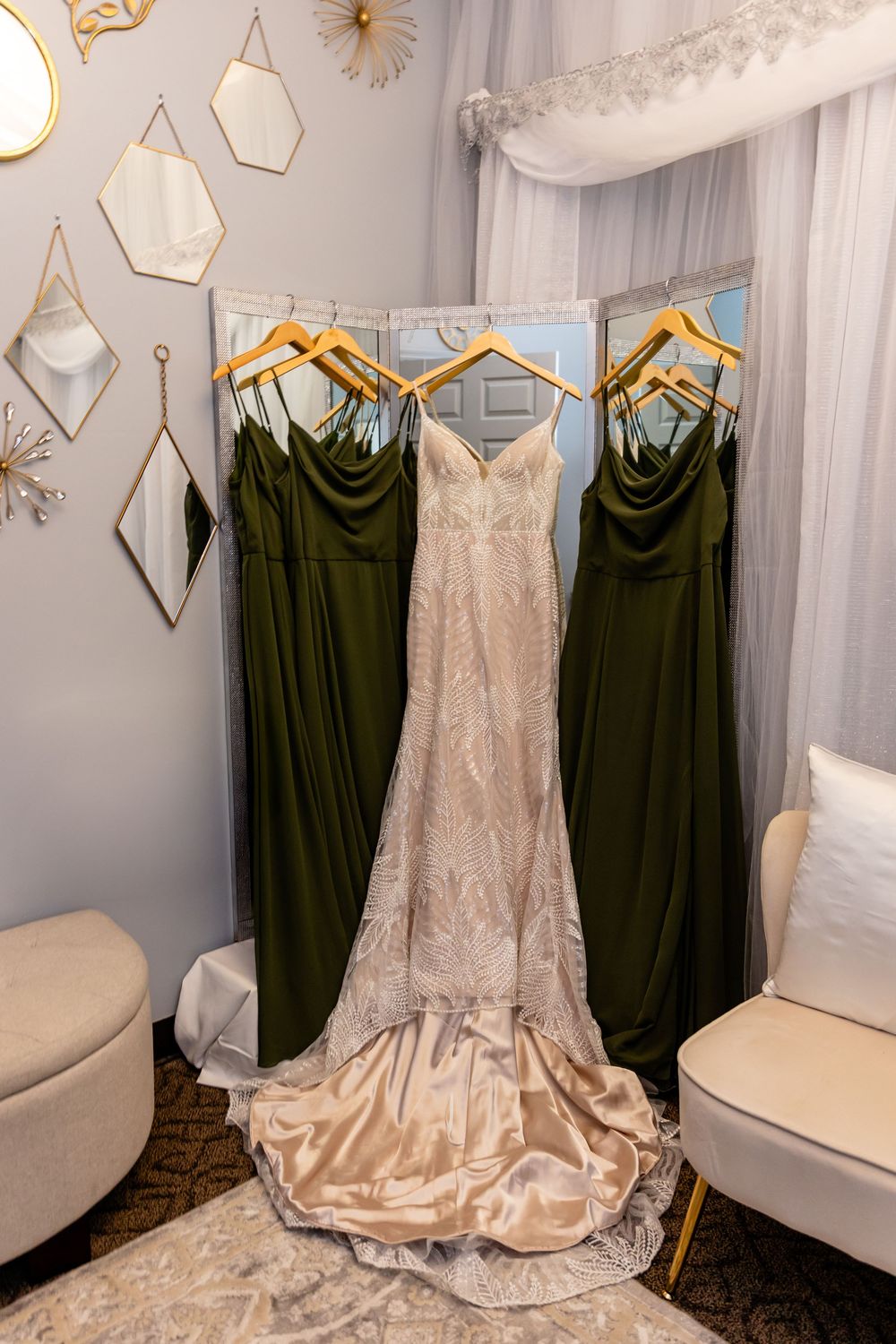 Elegant wedding dress with lace detail on wooden hanger beside olive green bridesmaid gowns in a bridal suite at a Venue on the Ridge