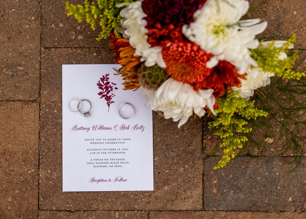 Wedding invitation with two rings beside autumn floral bouquet featuring red, orange, and white flowers on stone.