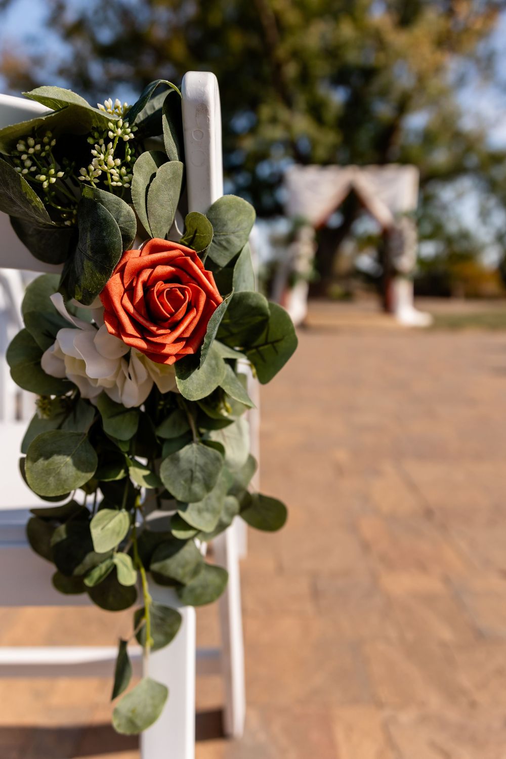 Orange rose and eucalyptus floral arrangement decorating white ceremony chair with outdoor wedding arch in background.