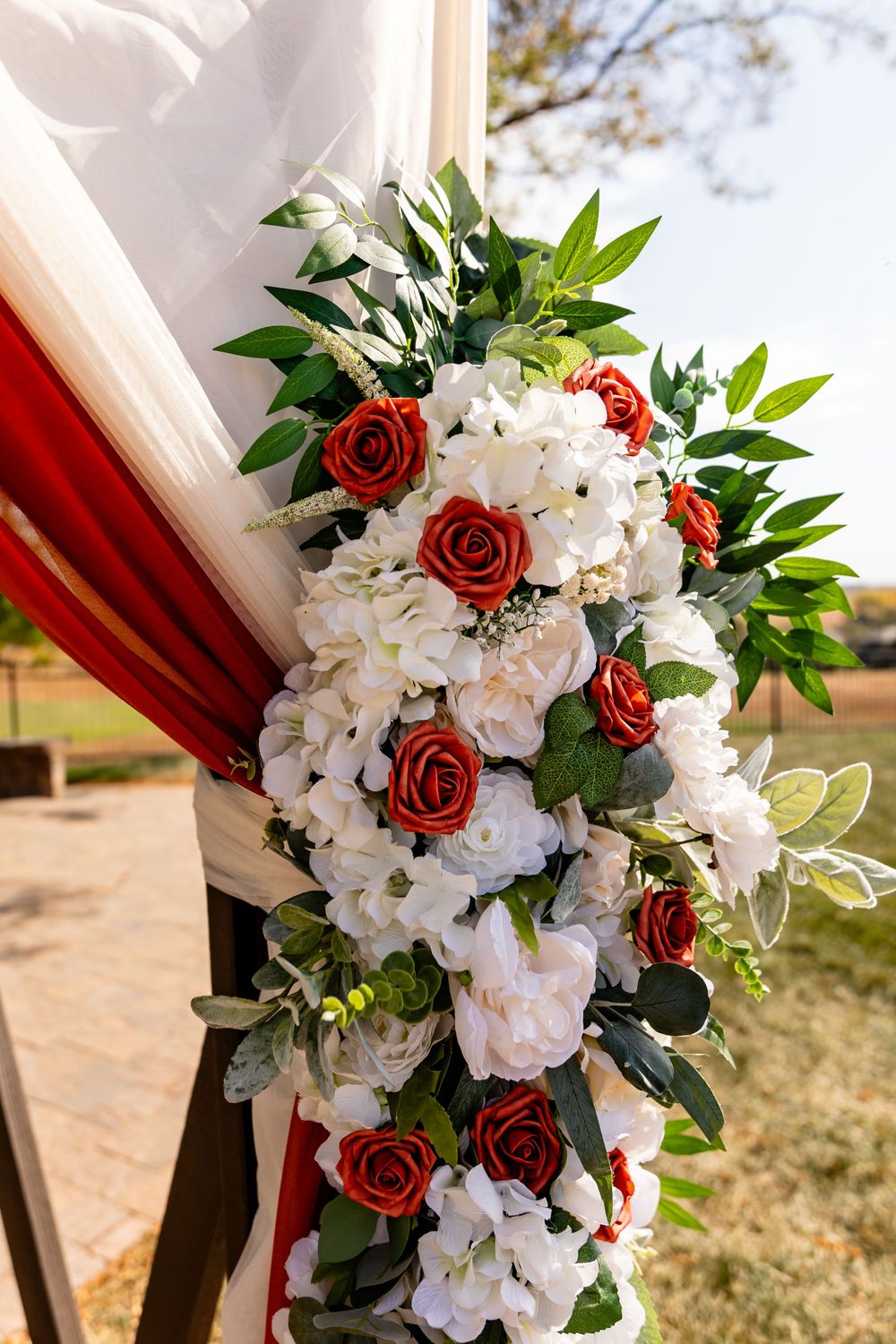 Red and white roses with green foliage adorn a wedding arch draped in white and red fabric outdoors.
