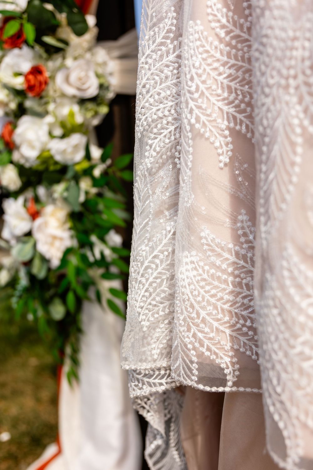 Close-up of a white lace wedding dress with intricate leaf embroidery next to a floral arrangement.