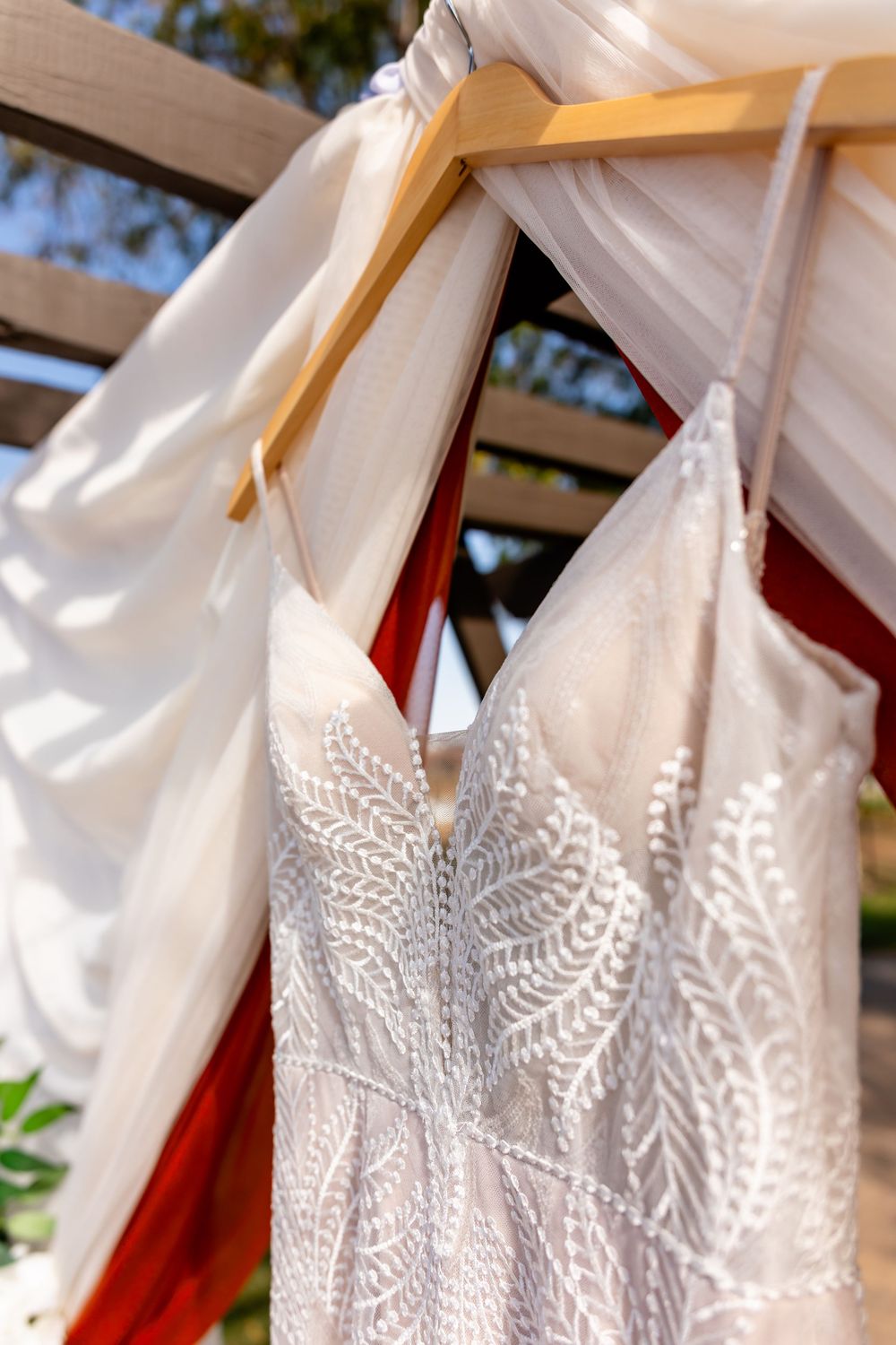 Elegant beaded spaghetti strap wedding dress hanging on a wooden hanger under a draped wedding arch.