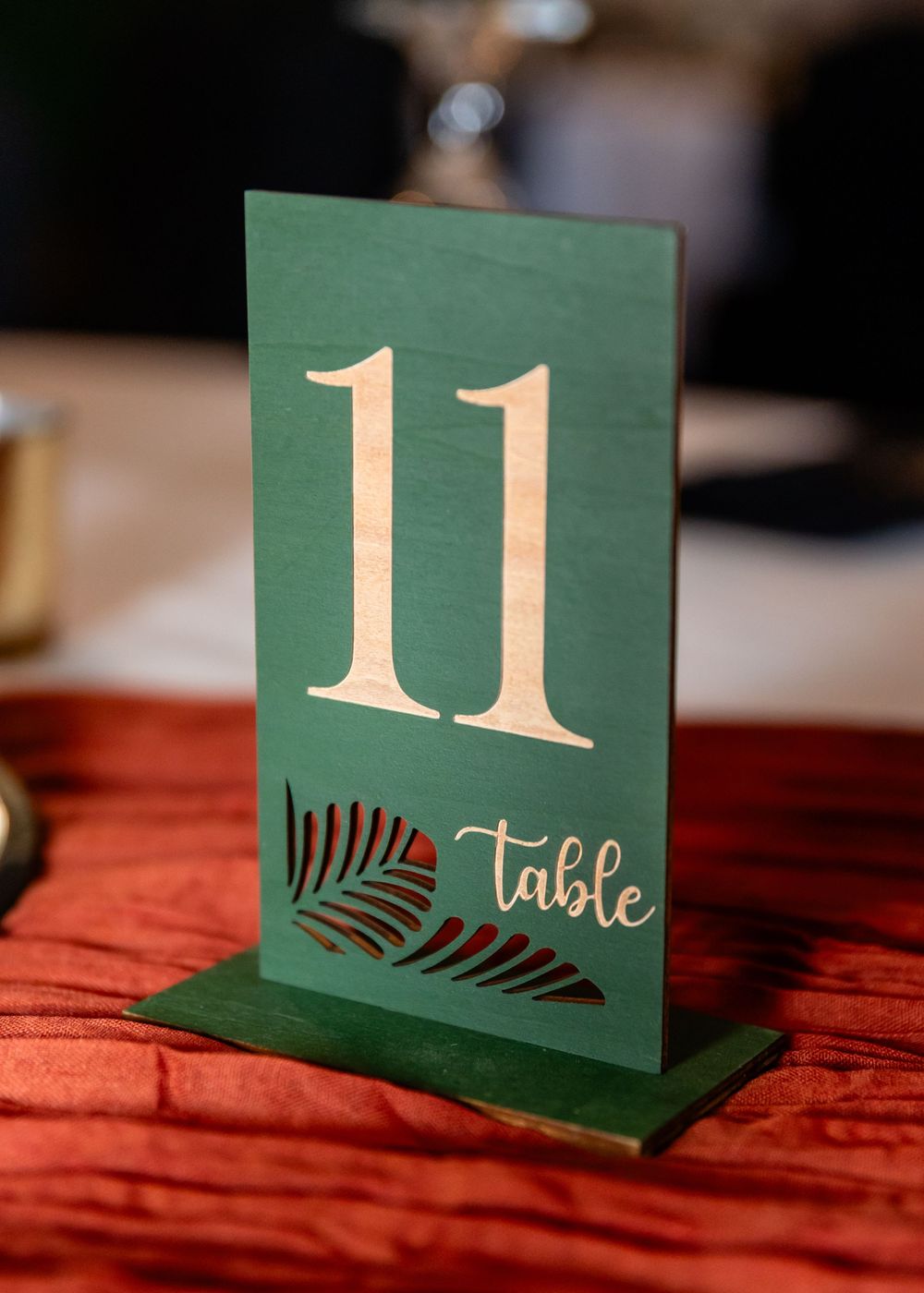 Green and gold laser-cut wooden table number 11 sign with palm leaf design on red satin tablecloth.