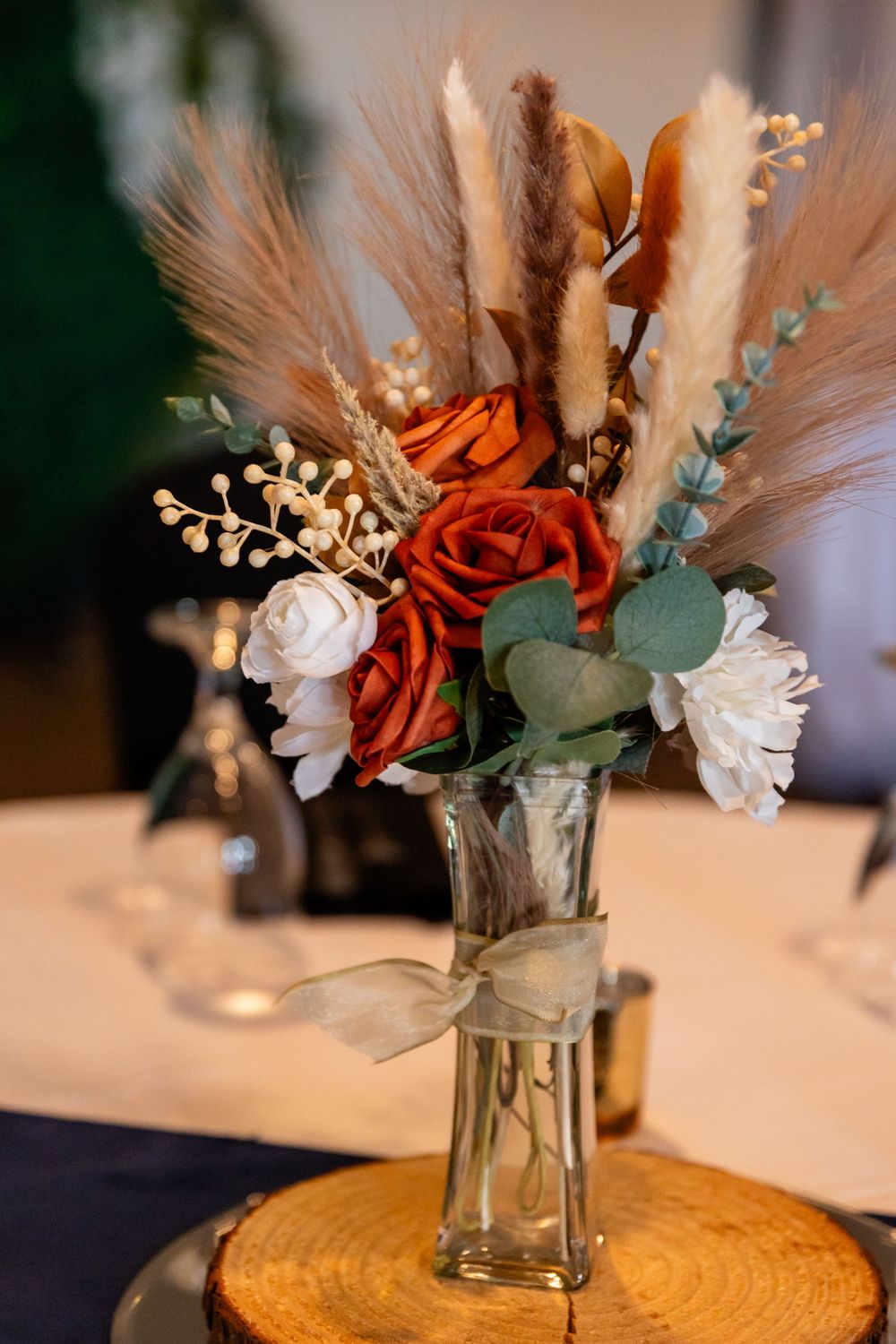 Rustic wedding centerpiece with red and white roses, pampas grass, and eucalyptus in a glass vase on a wood slice.