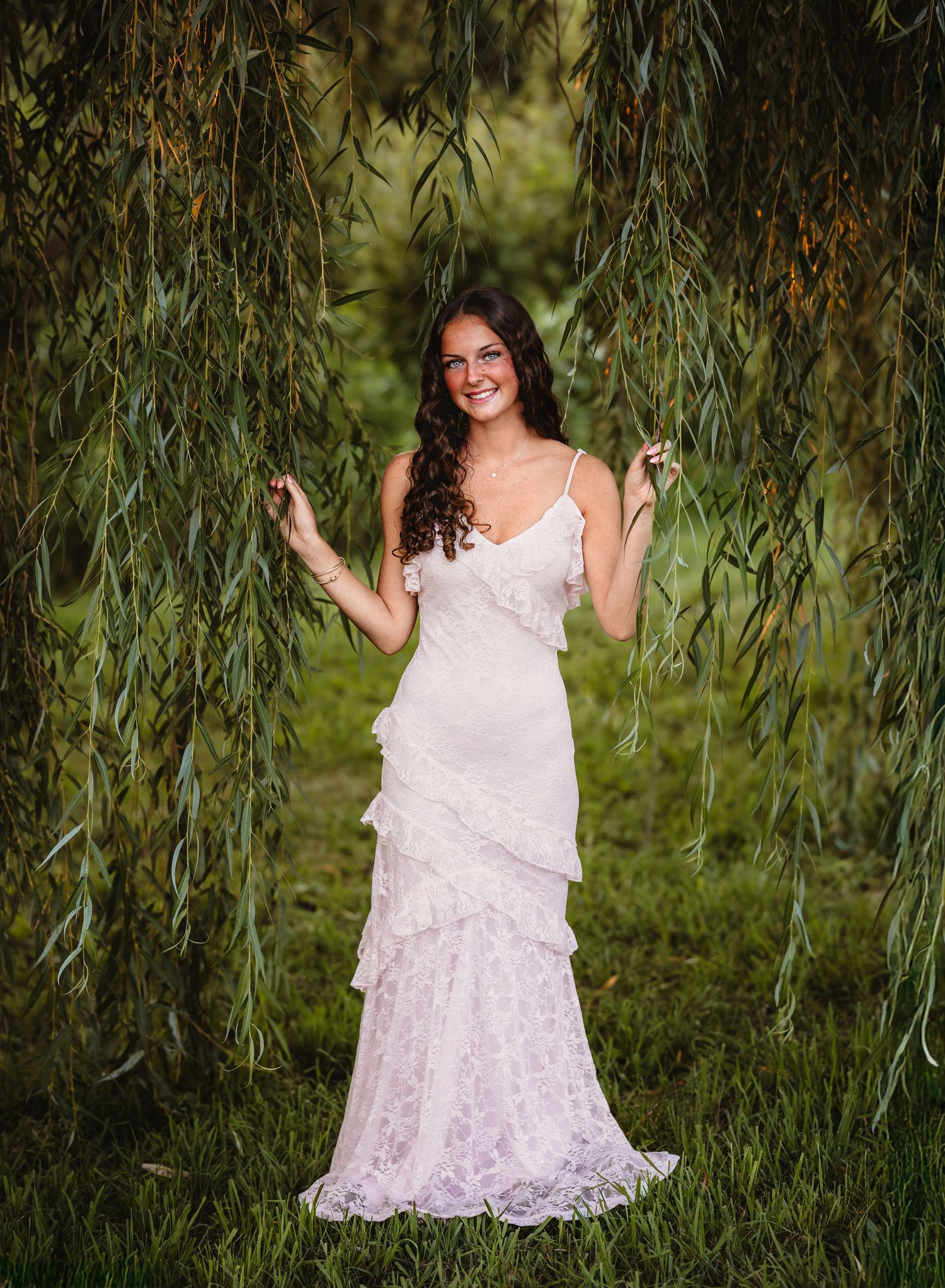 A senior girl in a pink lace dress poses gracefully among drooping willow branches in a natural outdoor setting.