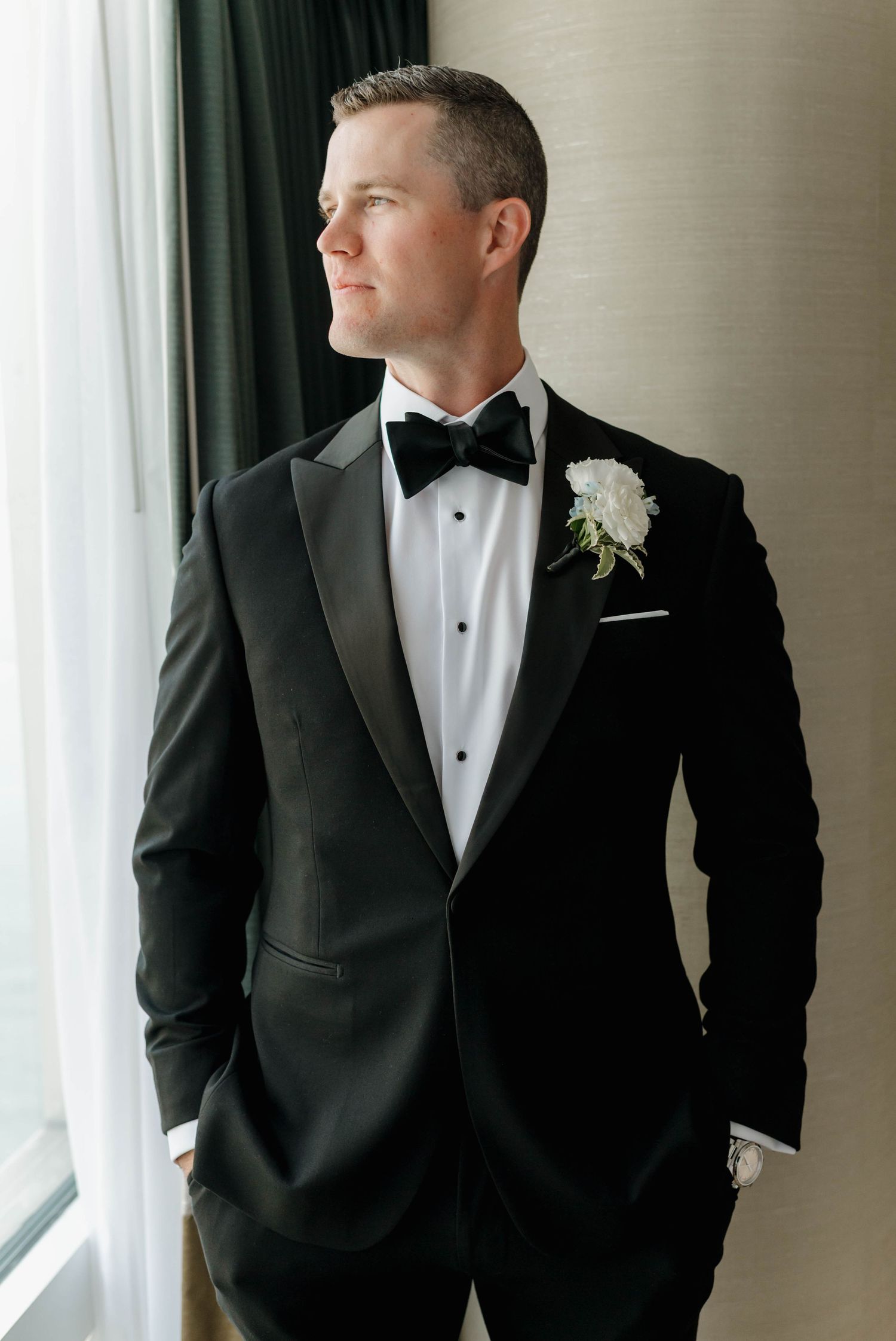 A groom in a classic black tuxedo and bow tie gazes thoughtfully out a window while getting ready for his wedding.