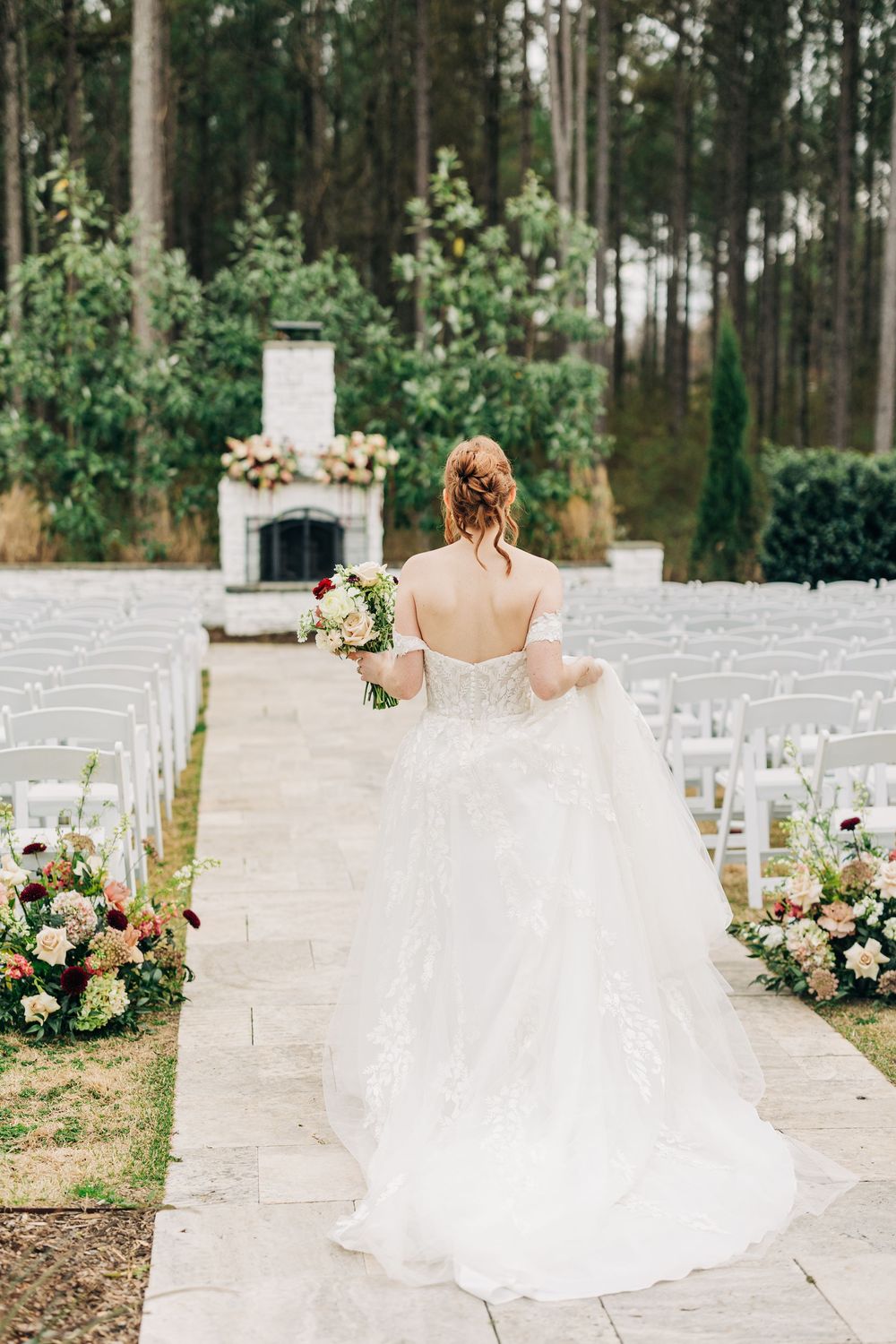A wedding ceremony setup in a wooded area with white chairs and floral arrangements leading to a stone fireplace altar.