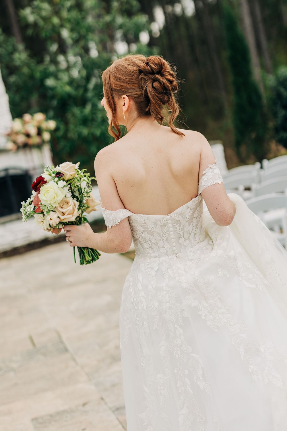 A bride in a white lace wedding dress holds a bouquet while walking through an outdoor garden setting.