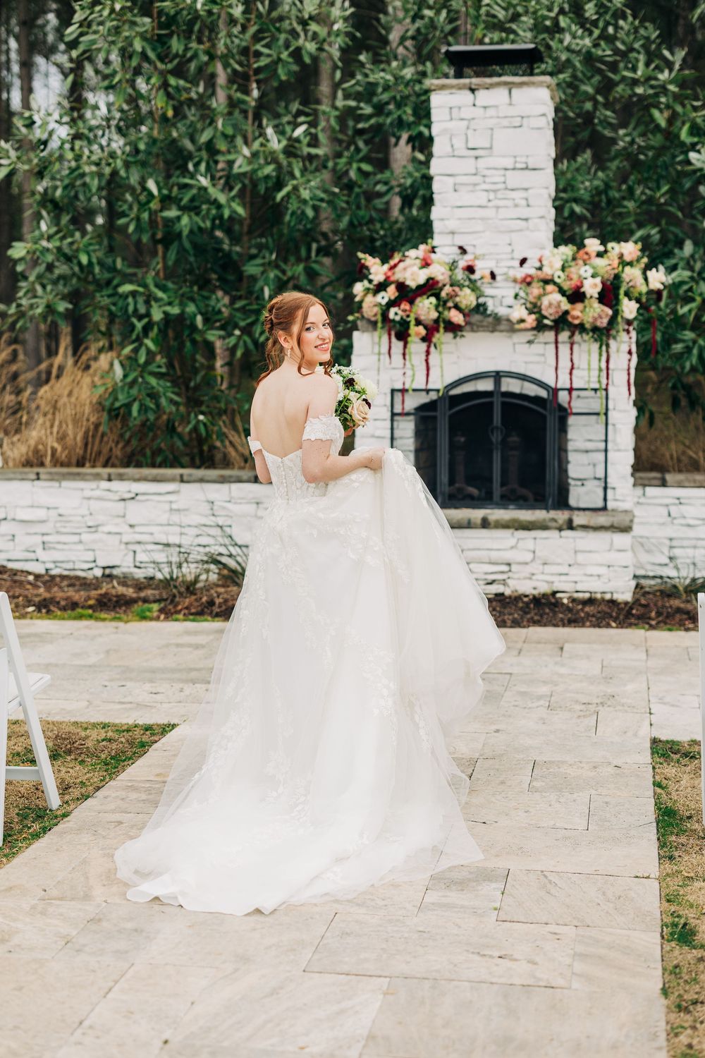 A bride in a flowing white gown walks along a stone path in an outdoor garden setting.