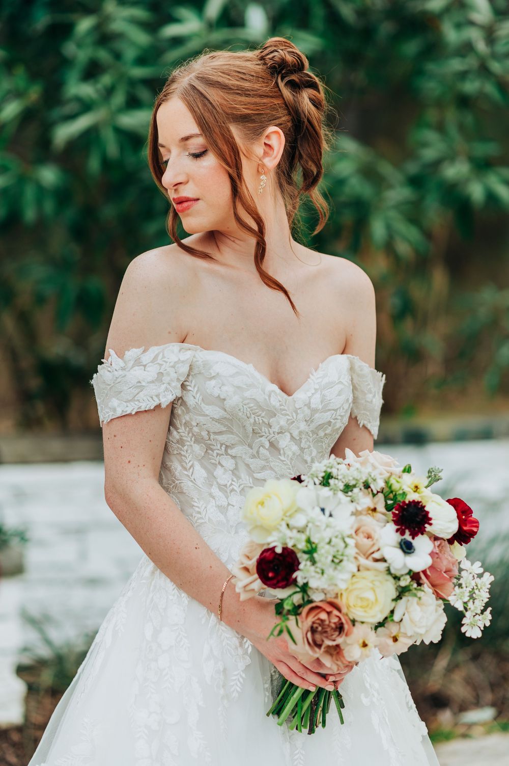 Close-up details of a bride in a white off-shoulder wedding dress holding a romantic bouquet with burgundy and cream flowers.