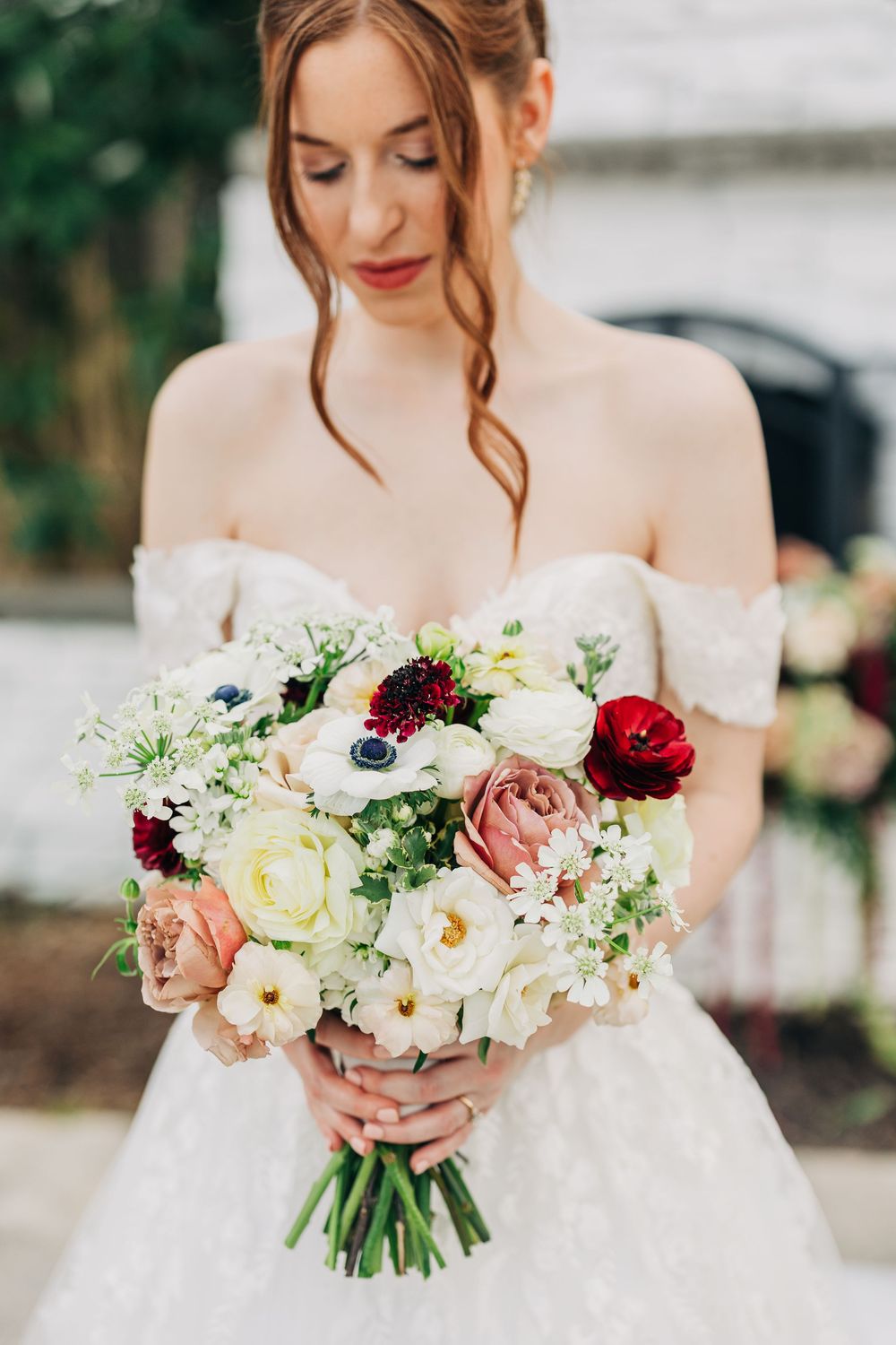 Elegant bridal bouquet features white anemones, blush roses, and deep red accents against an off-shoulder wedding dress.