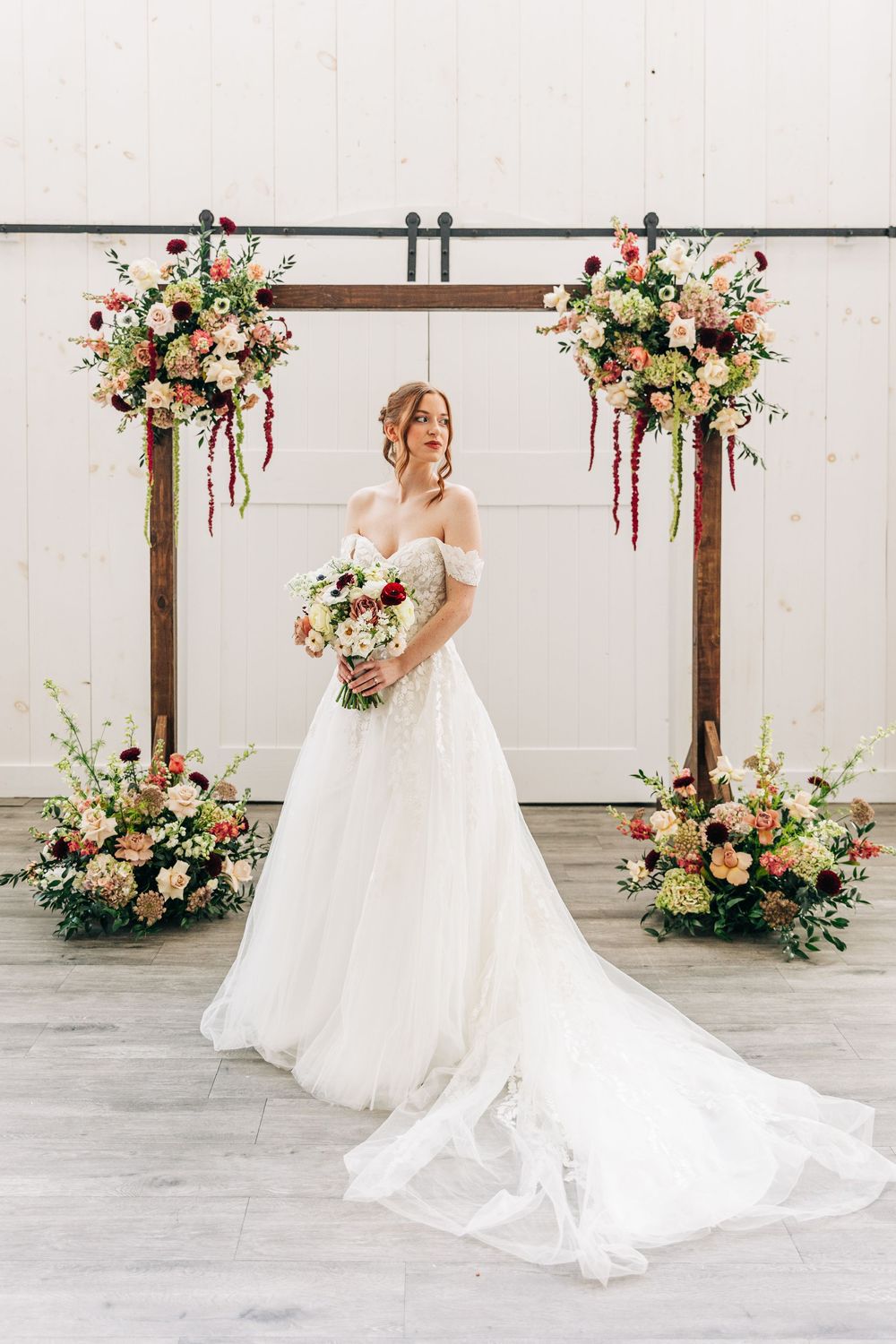 Elegant wedding arch decorated with peach and pink floral arrangements frames a bride in flowing off-shoulder gown.