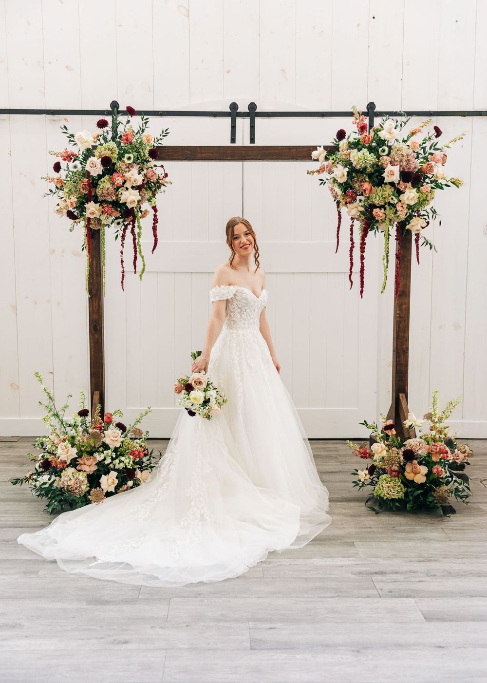 A bride in a flowing white gown stands in front of a rustic wooden arch adorned with peach and pink floral arrangements.