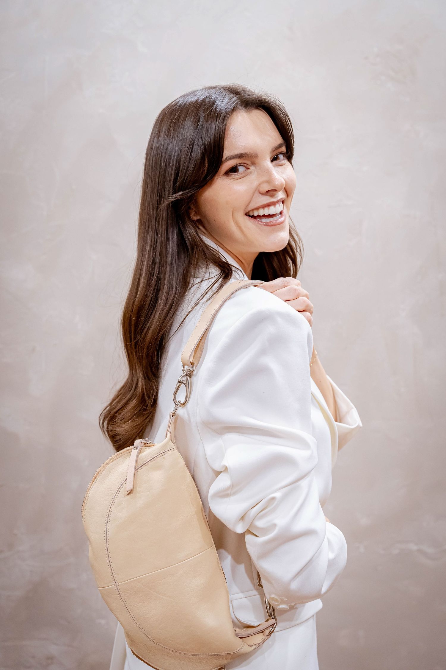 Smiling brunette woman wearing white outfit with beige leather backpack over shoulder against neutral background.