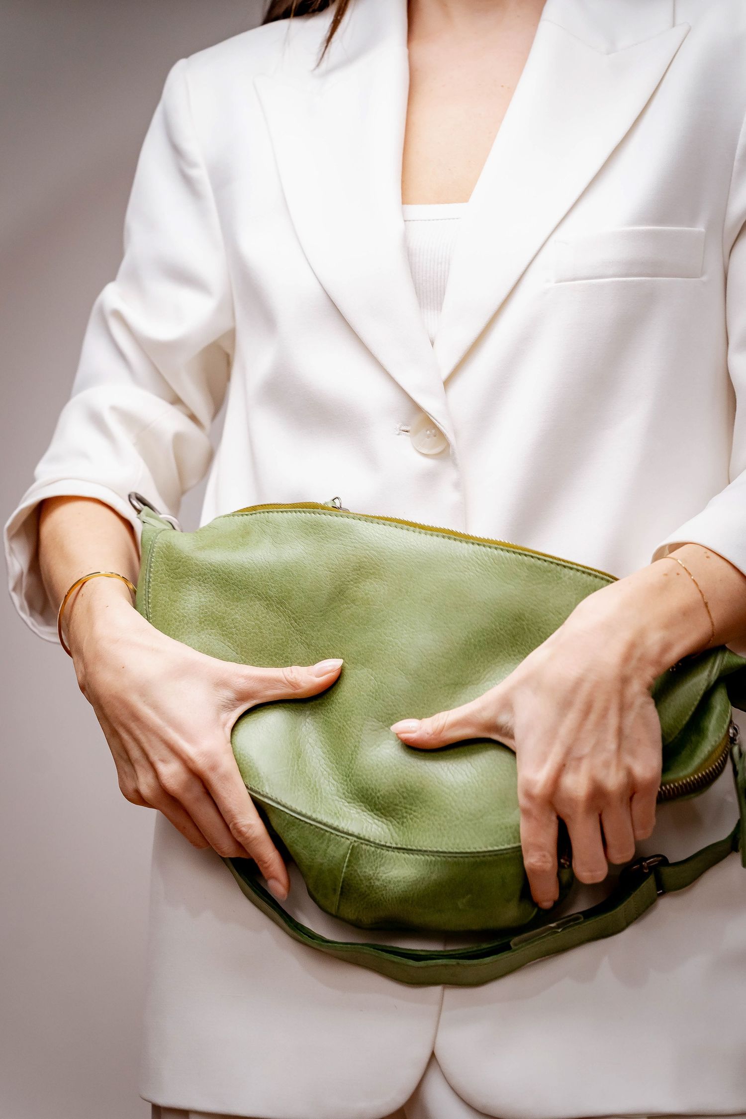 A woman in a white blazer holding a green pebbled leather crescent sling bag with both hands.