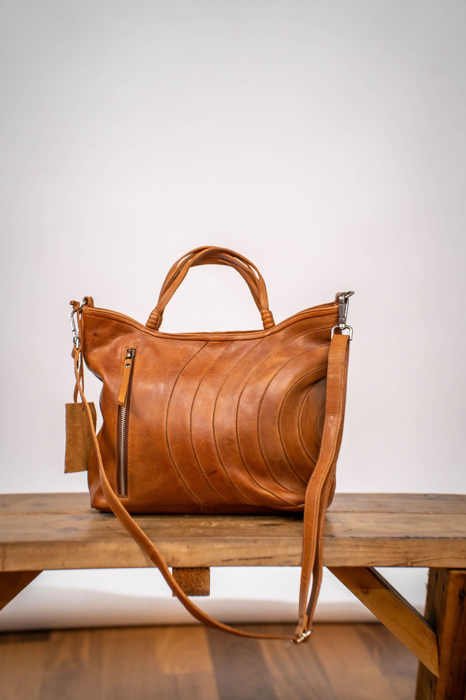 Tan brown leather tote bag with curved stitching detail and crossbody strap on wooden bench.