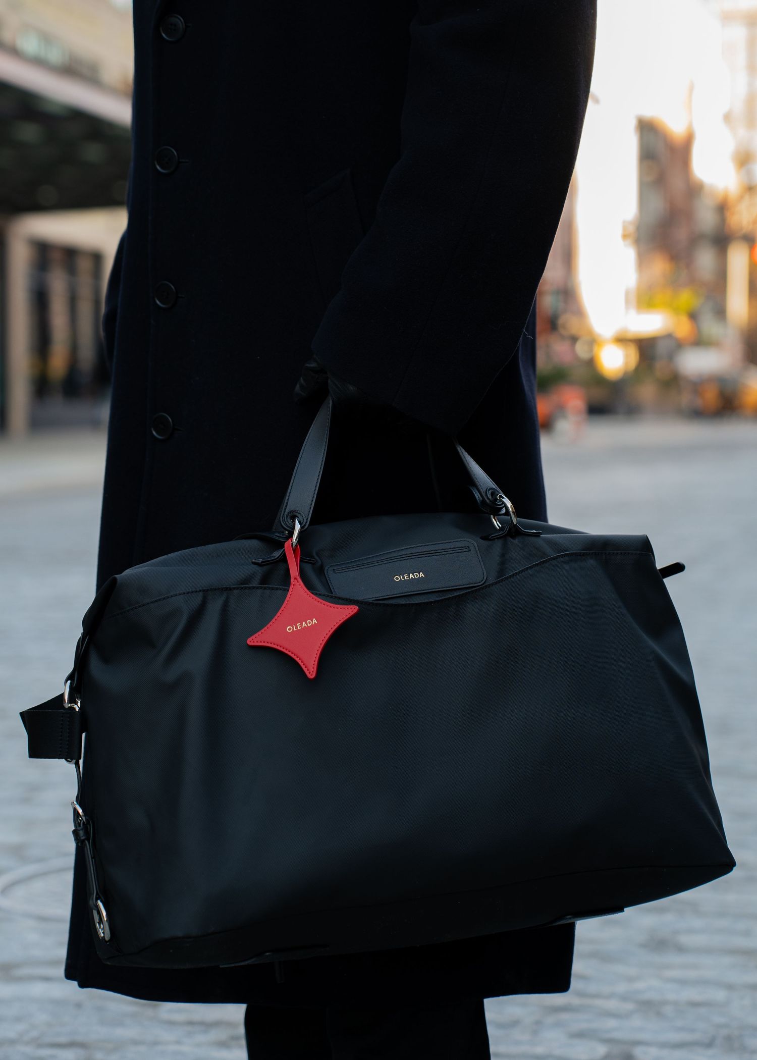 Person in black coat carrying a black nylon duffle bag with a red star-shaped luggage tag outdoors.