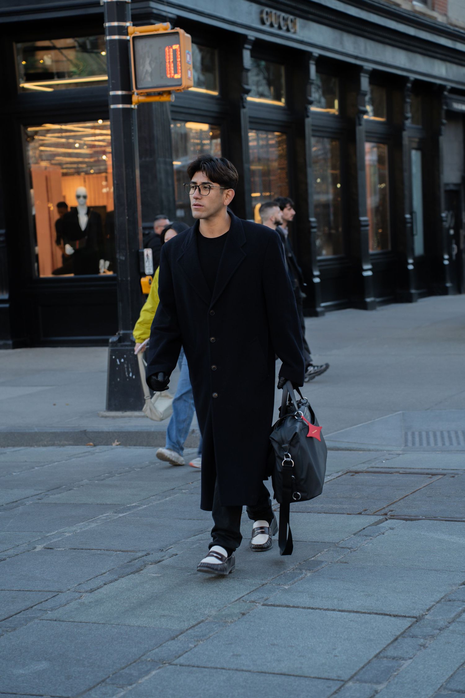 Asian man in long black overcoat and glasses walking on NYC sidewalk carrying black duffle bag.