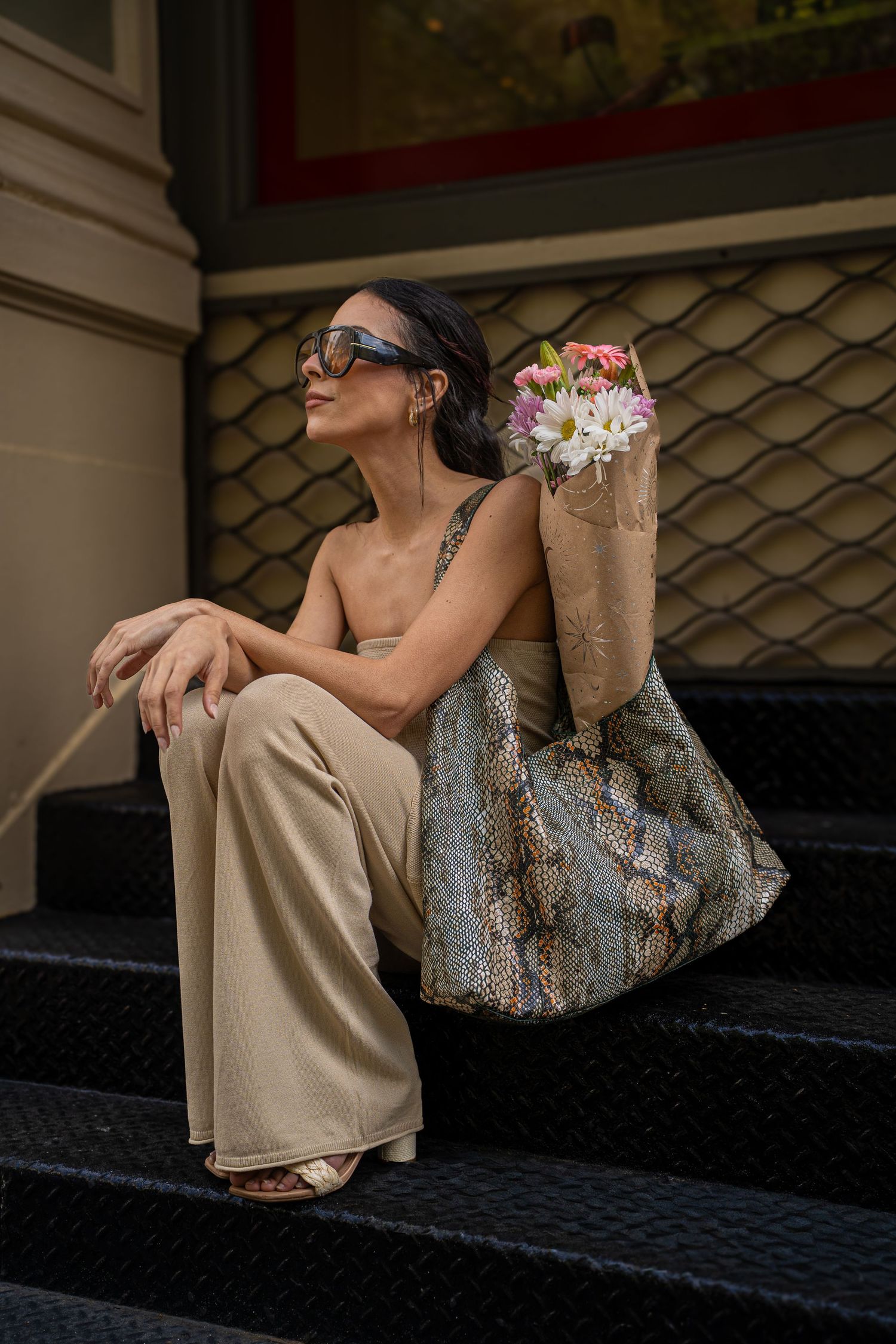 Woman in beige wide-leg pants and sunglasses sits on steps holding a floral bouquet in a snakeskin tote bag.