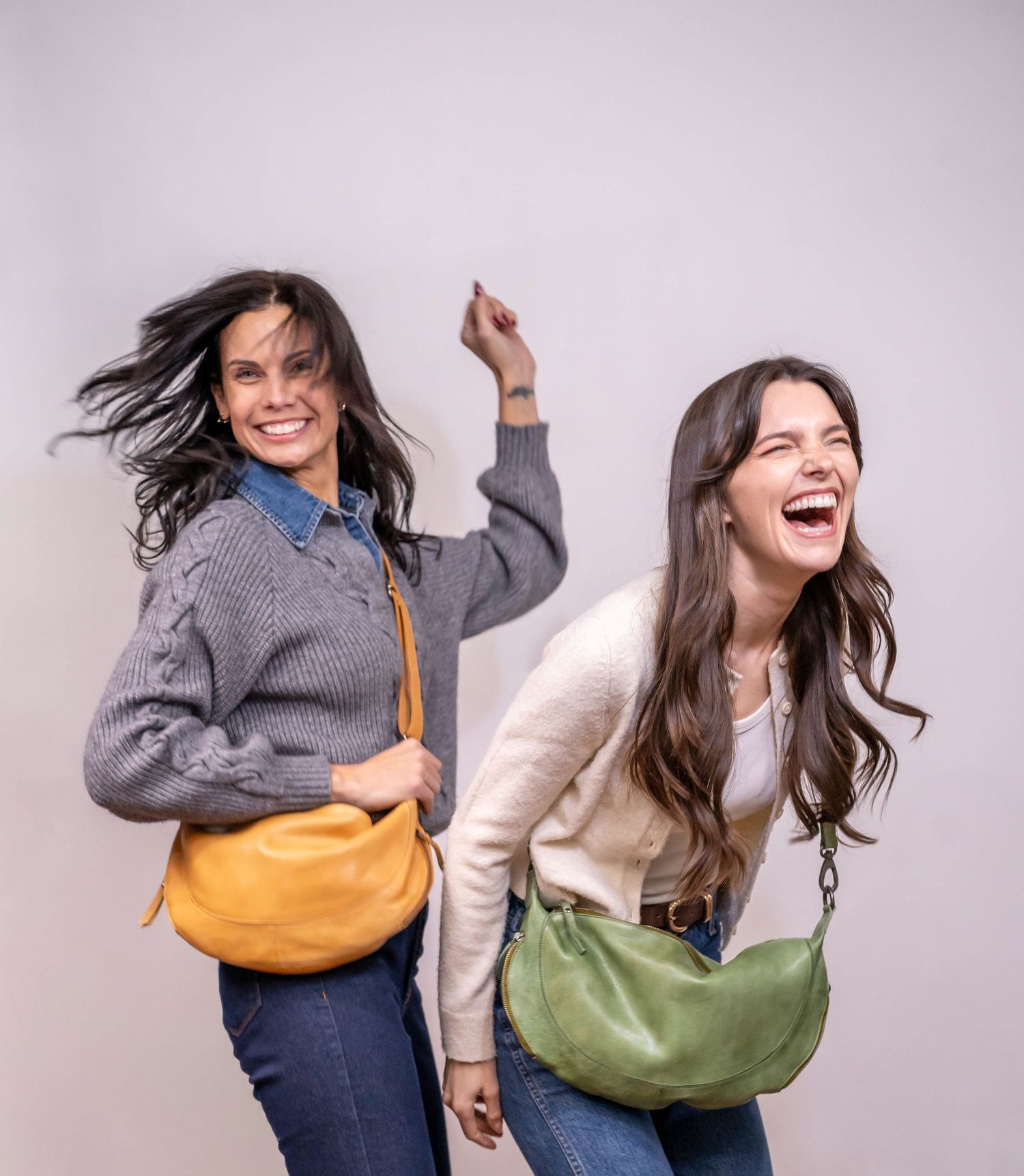 Two women laughing and posing with yellow and green leather crossbody bags.
