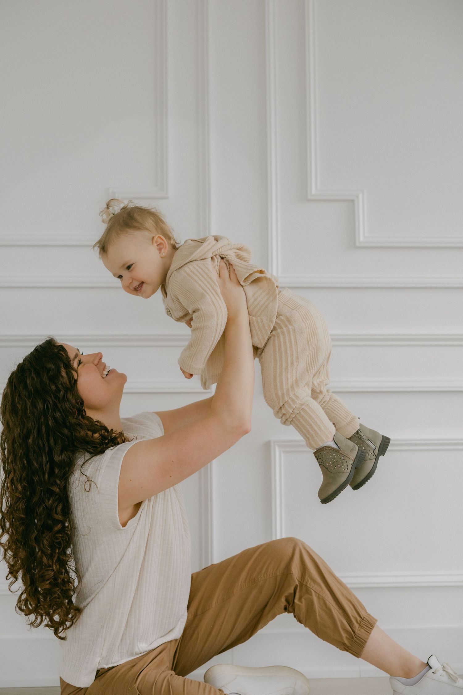 A parent lifts a small child wearing beige overalls while playing together against a white paneled wall.