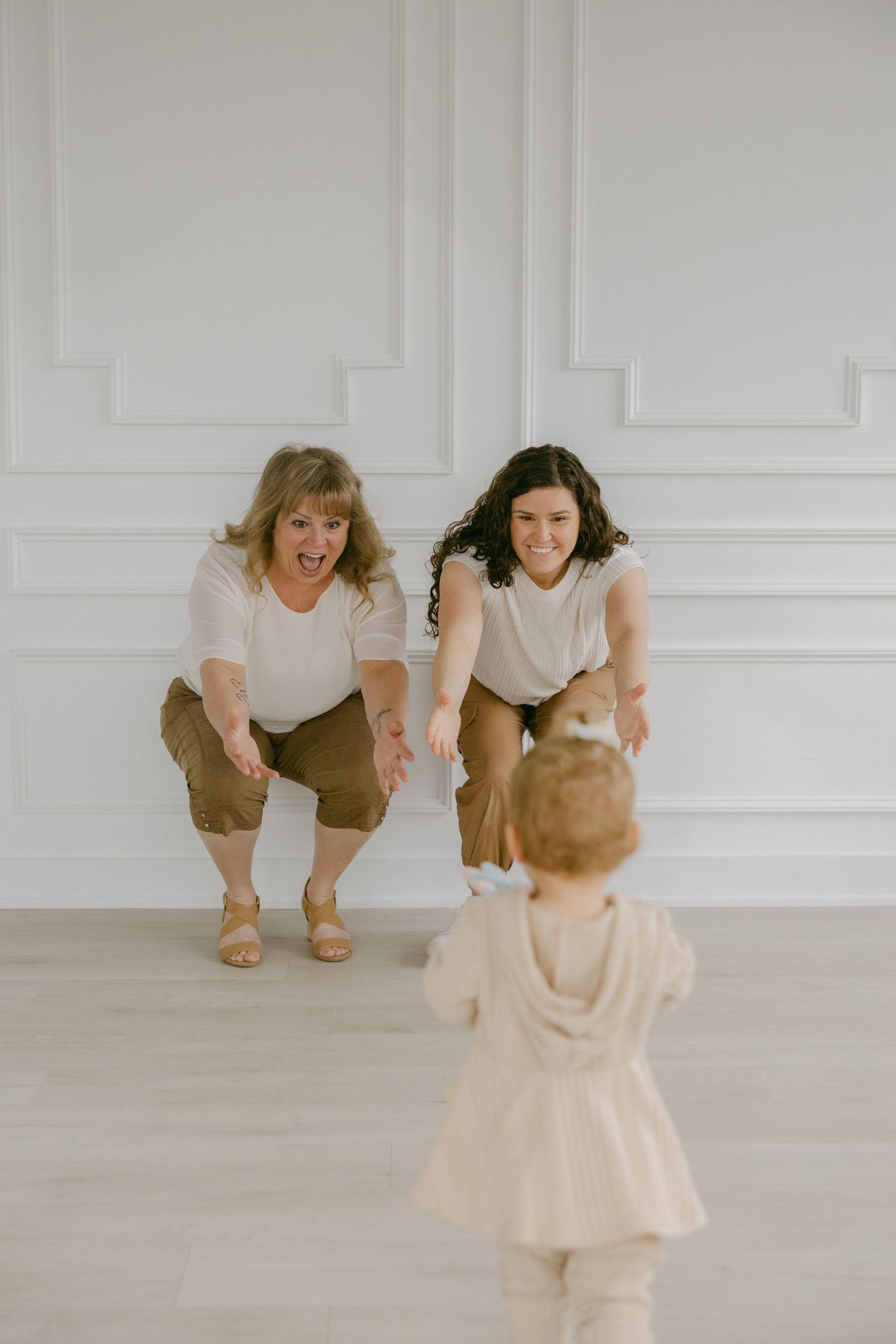 Two people in light colored outfits crouch down to interact with a small child in a bright room.