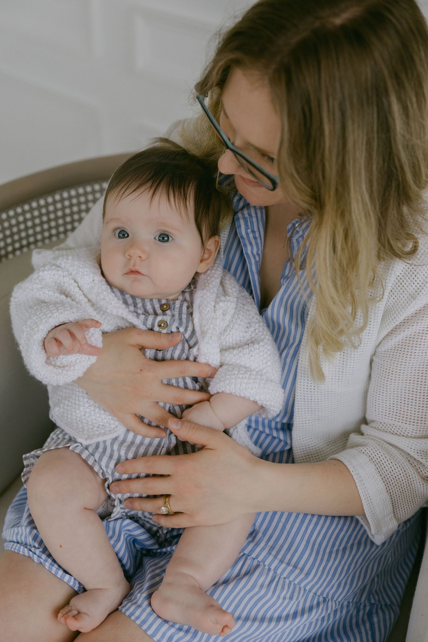 A baby in a white outfit sits in someone's lap while wearing a blue and white striped romper.