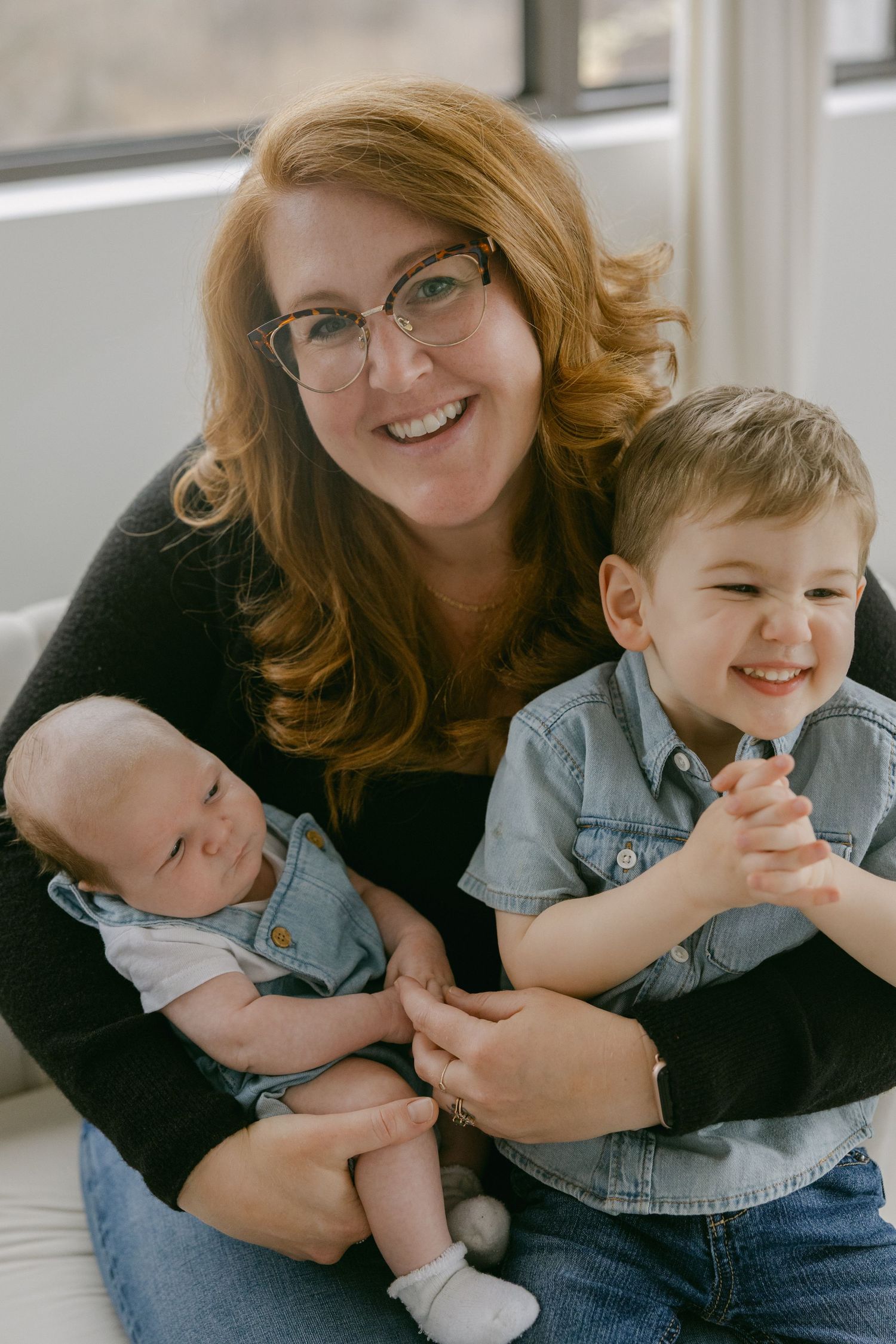 A family portrait showing a parent with two children, one baby and one toddler, sitting together by a window.