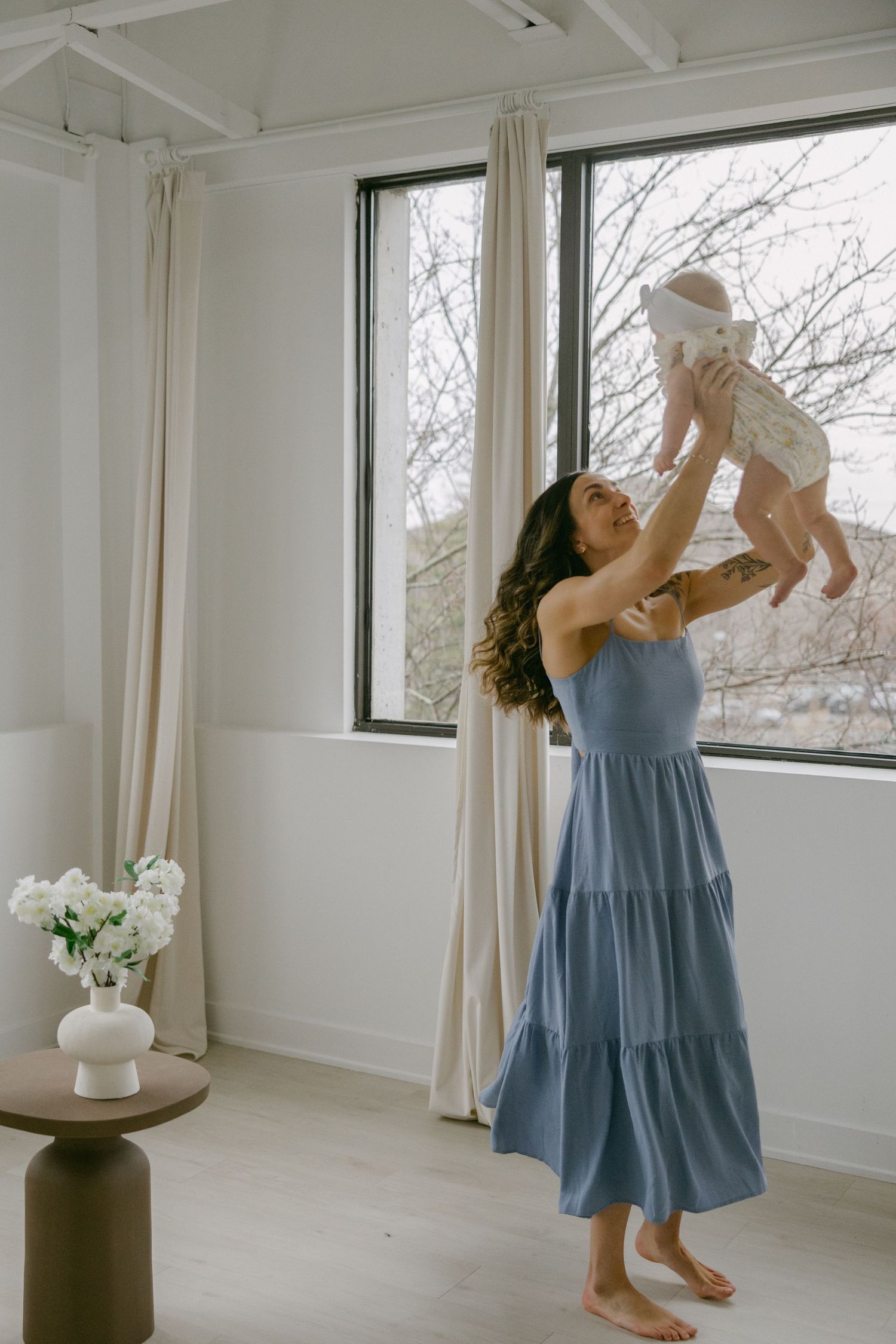 A woman in a flowing blue tiered dress lifts a baby in a bright sunlit room with white walls and large windows.