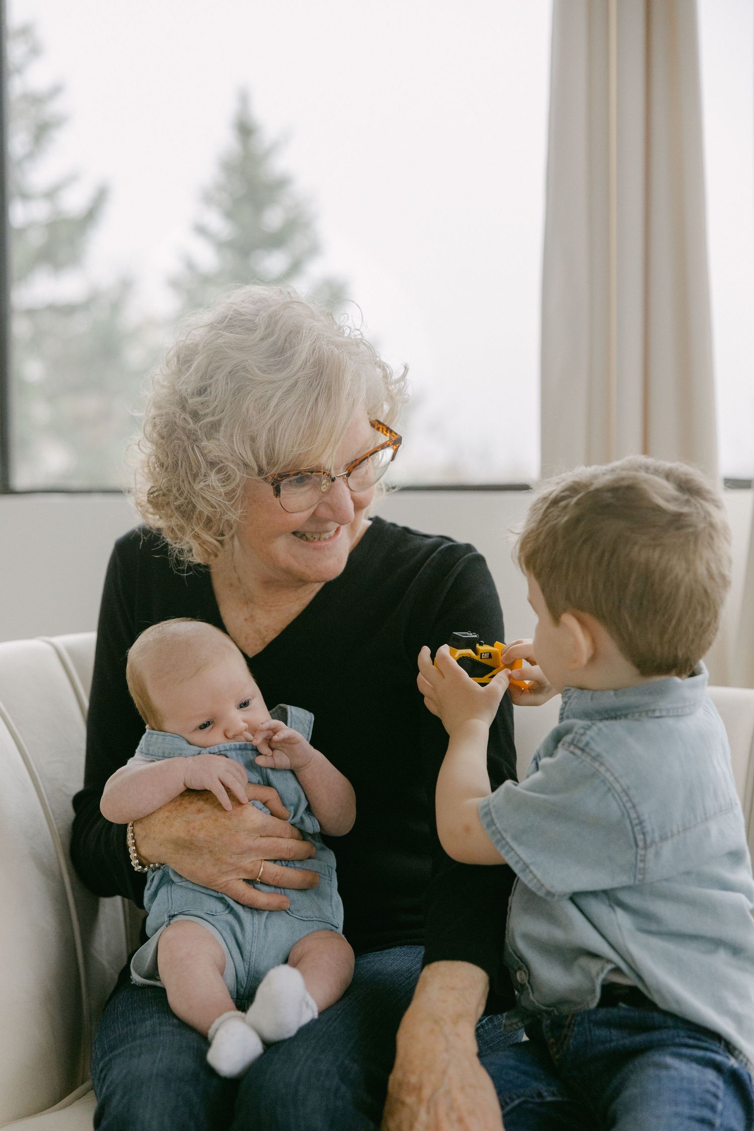 A tender family moment on a couch as a baby and young child spend time with an older woman in glasses by a window.