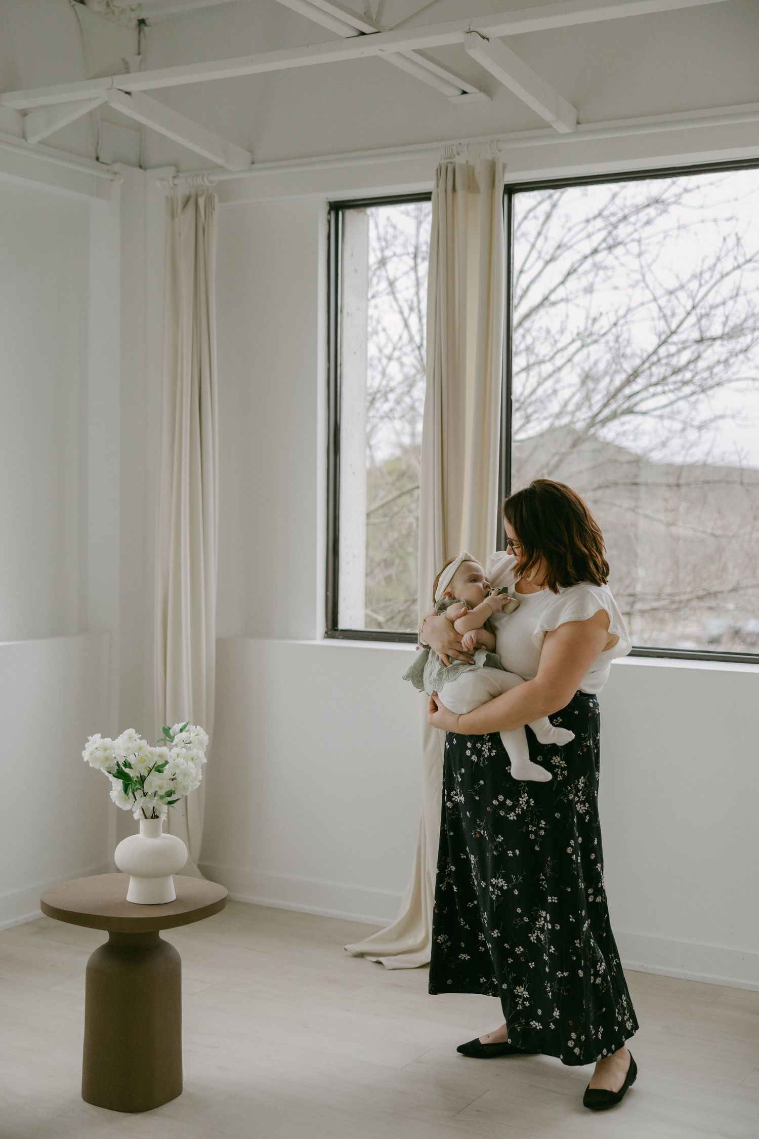 A person in a black floral dress poses elegantly near bright windows in a minimalist white studio space.