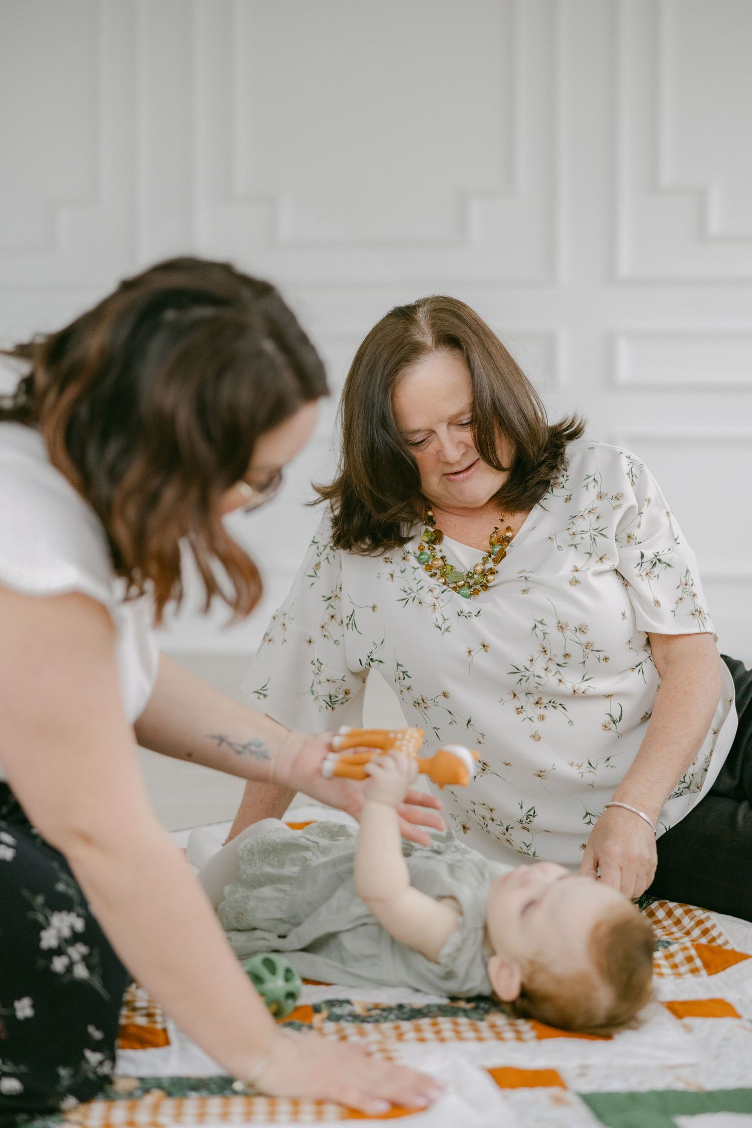 Two people sit on a colorful patchwork blanket with a baby, sharing drinks and enjoying a casual indoor gathering.