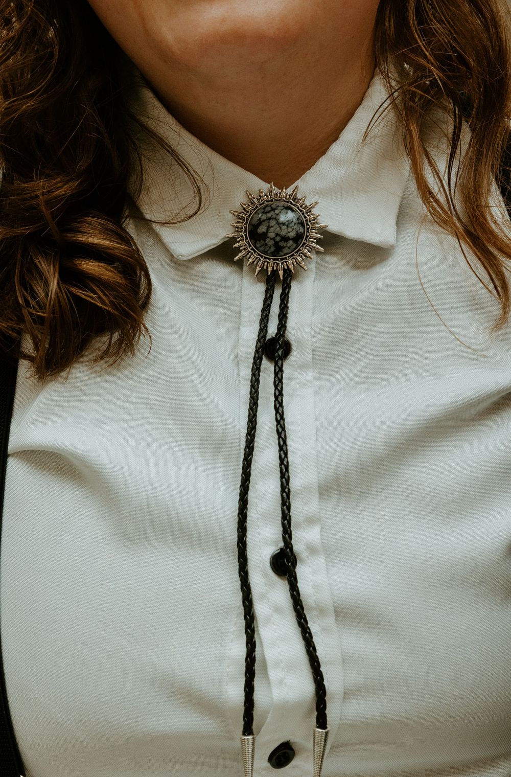 Woman wearing a silver sunburst bolo tie with dark stone on a white button-up shirt.