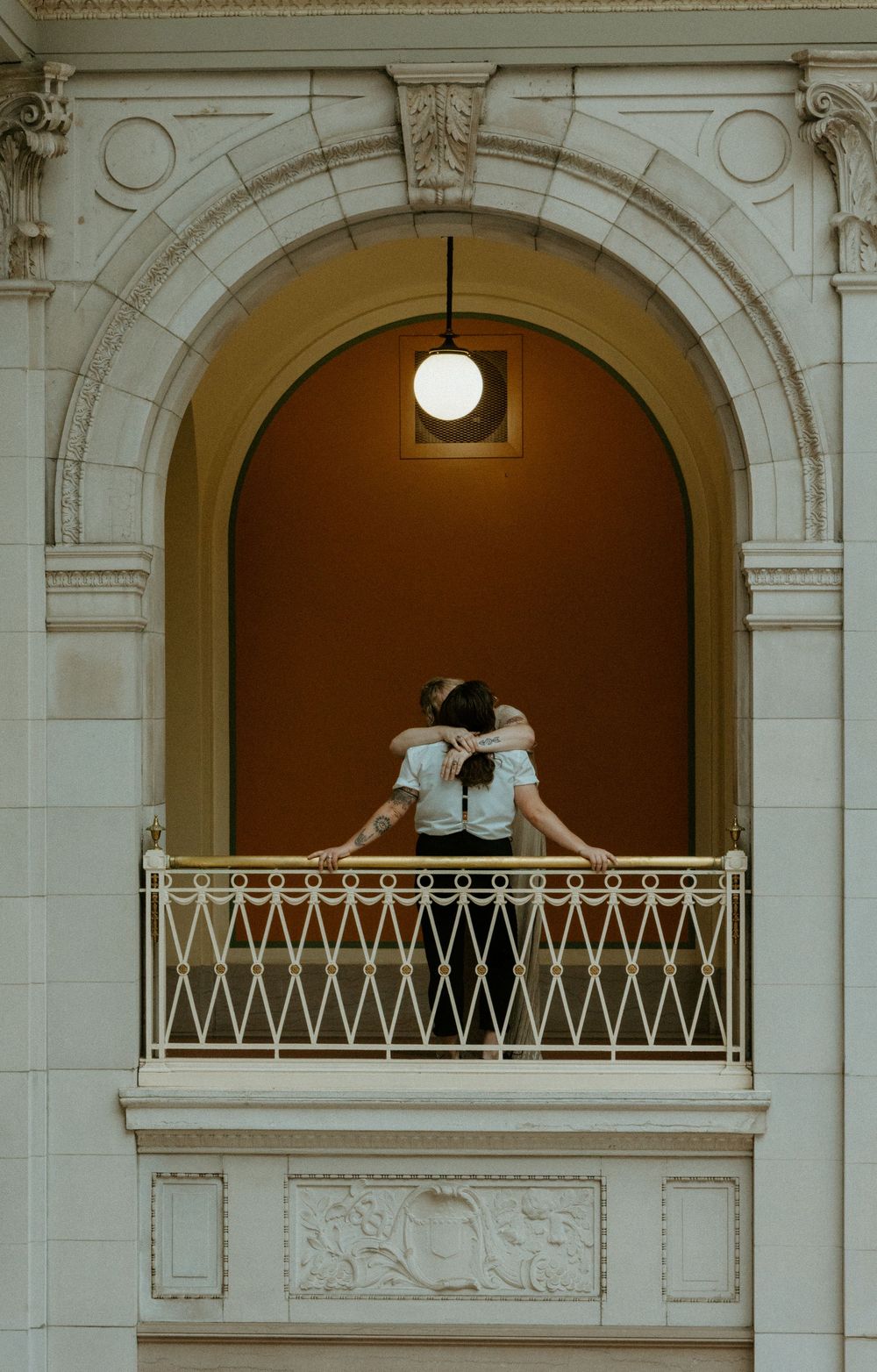 Couple embracing on ornate balcony with iron railing inside elegant arched architectural niche with globe light.