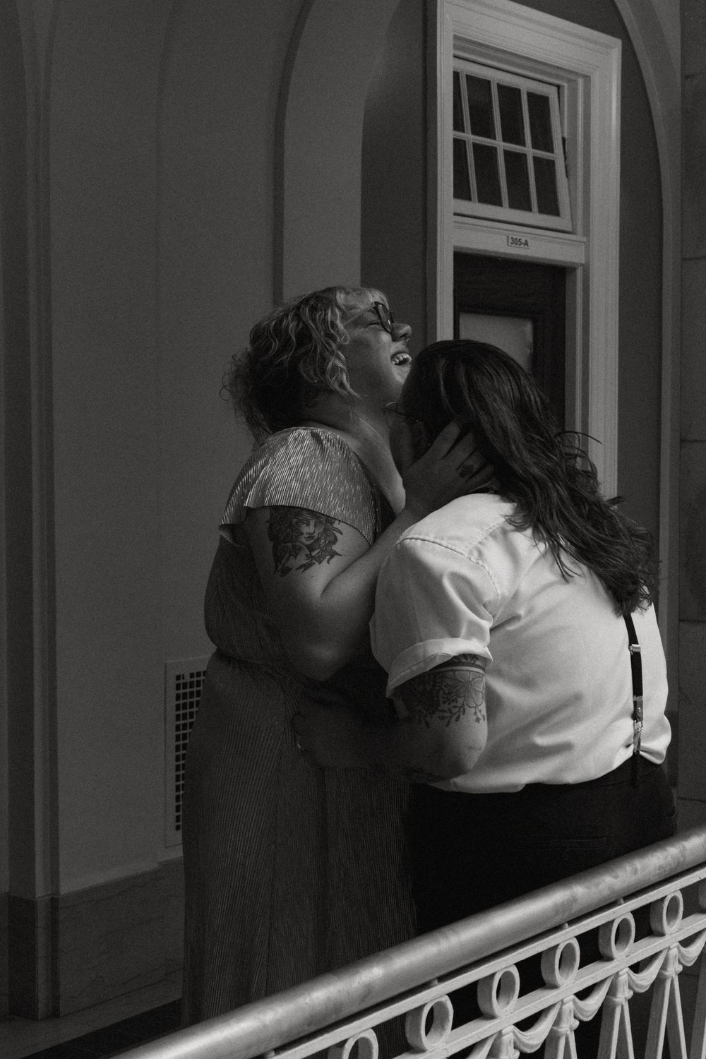 Black and white photo of two women laughing and embracing on a balcony, one in a lace dress and the other in suspenders.