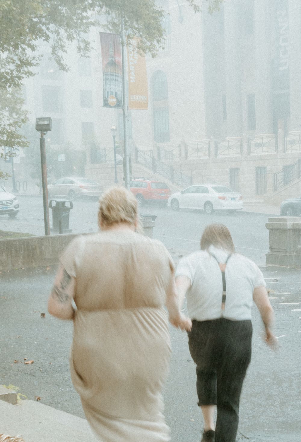 Two people walking through a rainy urban street with buildings and cars visible in the background.