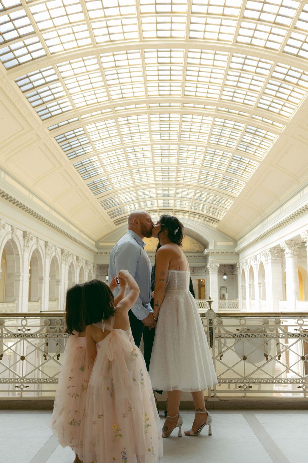 Couple sharing a kiss at an elegant museum wedding venue with a glass ceiling, while a flower girl twirls nearby.