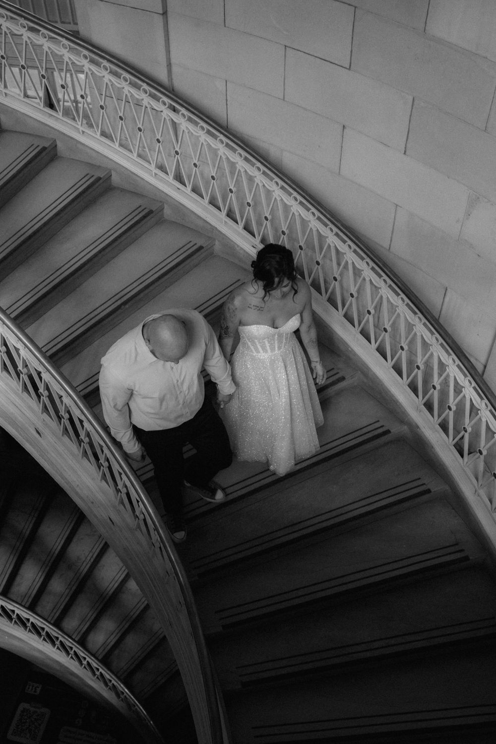 Bride and groom holding hands on elegant curved staircase, black and white wedding photo shot from above.