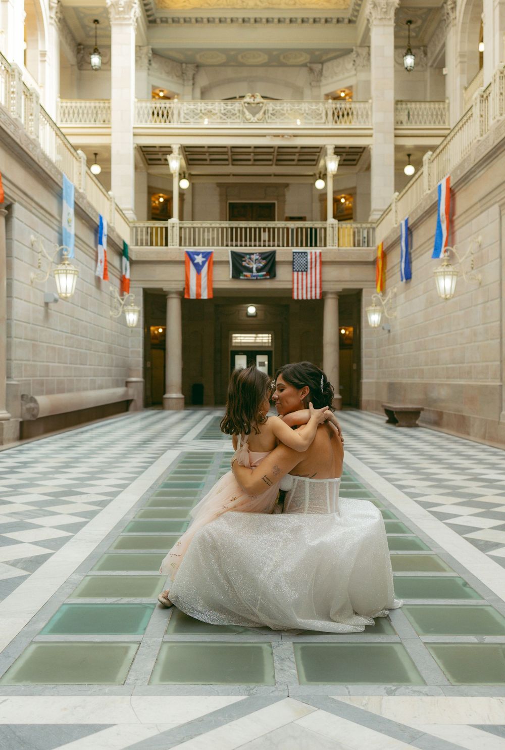 A bride and flower girl share a sweet hug in elegant grand hall adorned with international flags.