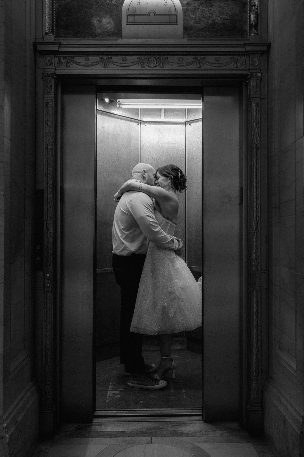 Black and white photo of a couple embracing and kissing inside an elegant elevator, bride in white dress.