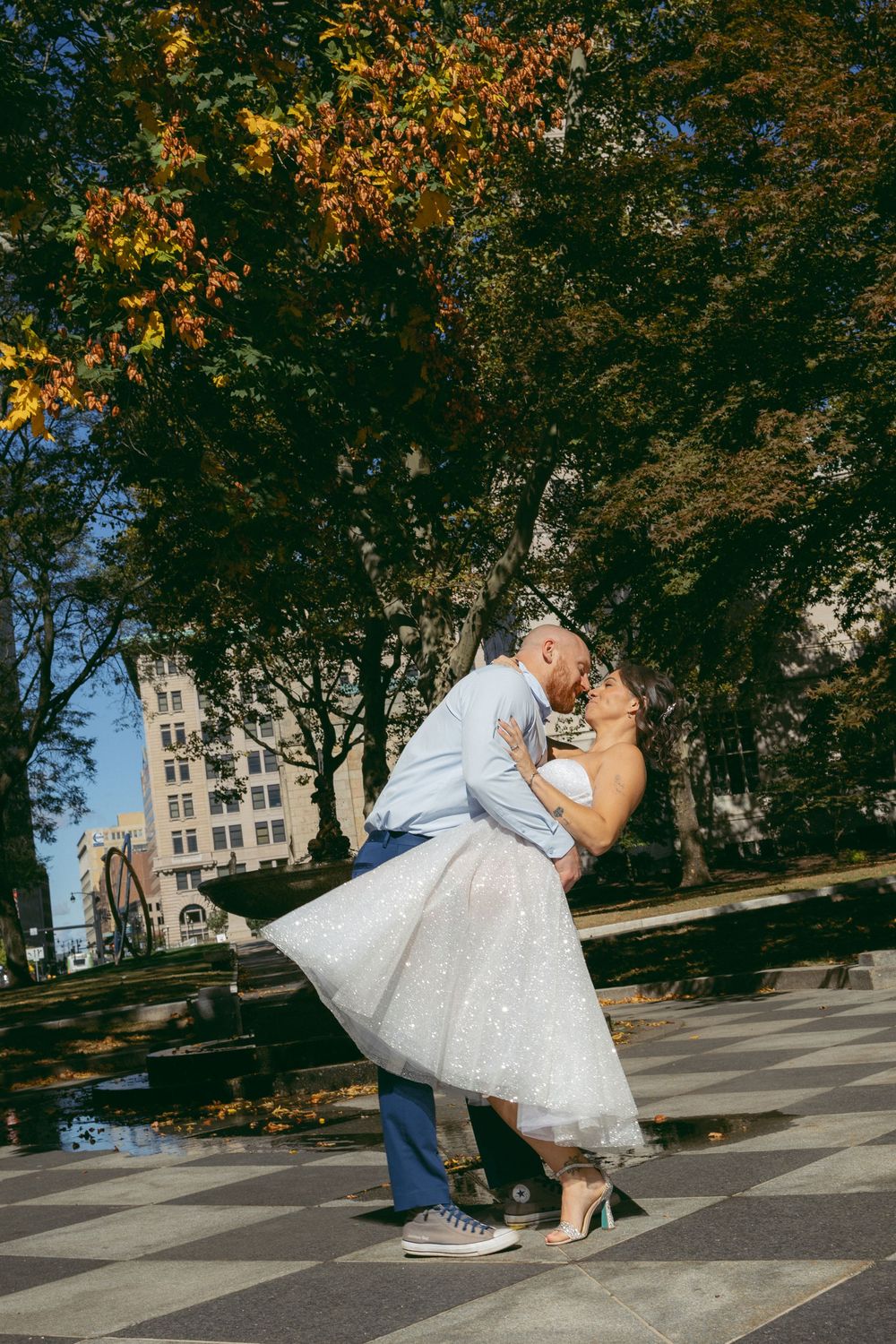 Groom dips bride in white tea-length dress for a kiss outdoors near a fountain with autumn trees in an urban park.