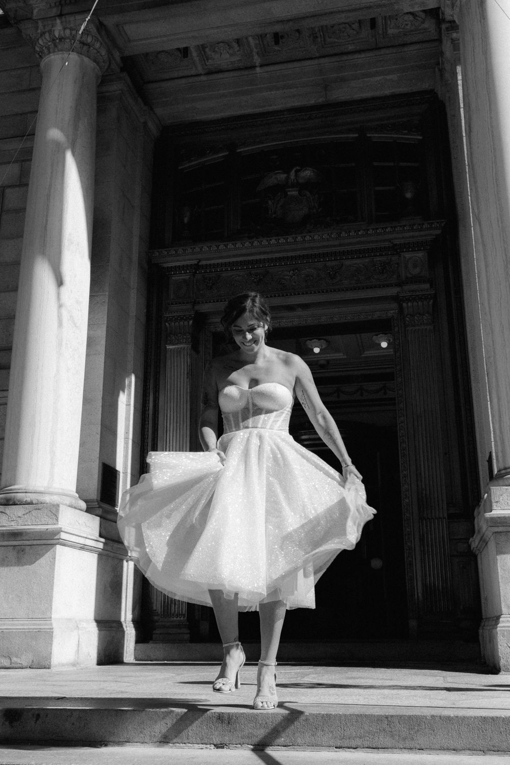 Black and white photo of a woman in a strapless white tulle dress posing on steps of a grand classical building.
